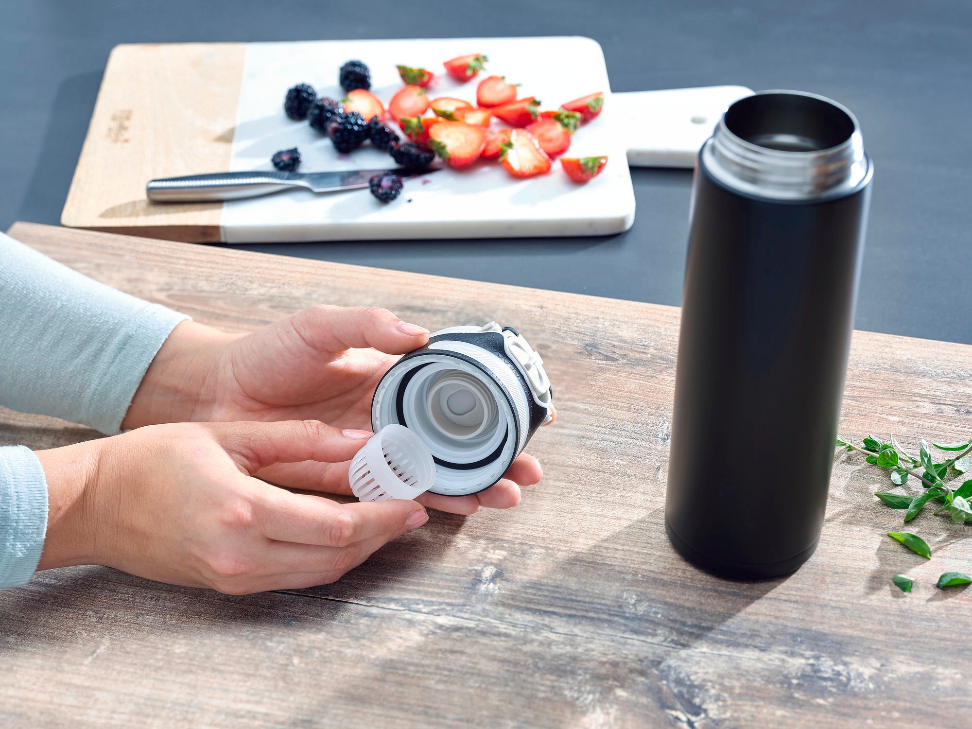 Flip 600 ml insulated mug in black on a table. A person is holding the lid with integrated strainer. In the background is a chopping board with sliced fruit.