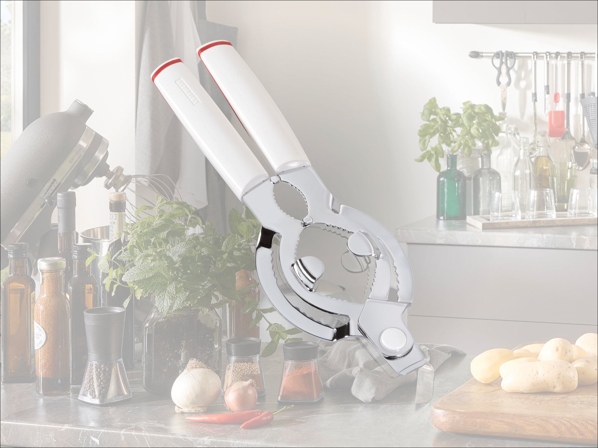 A high-quality bottle opener with ergonomic white handles rests on a modern kitchen worktop. Herbs, spices and kitchen utensils can be seen in the background.