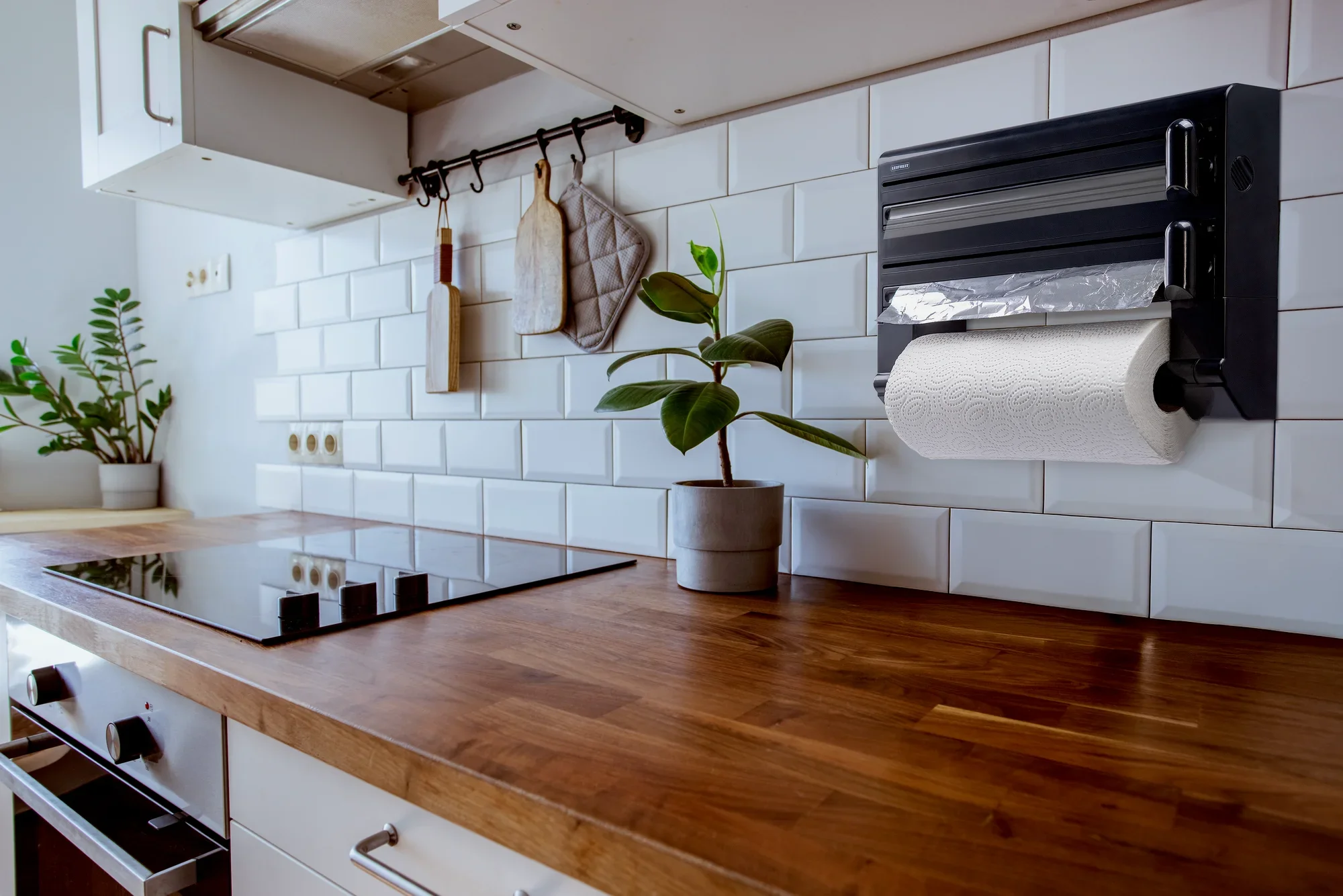 Wandrollenhalter Parat Black von Leifheit in einer modernen Küche. An der weißen Fliesenwand montiert, bietet er Platz für Folien und Küchenpapier. Daneben Pflanzen und Küchenutensilien.
