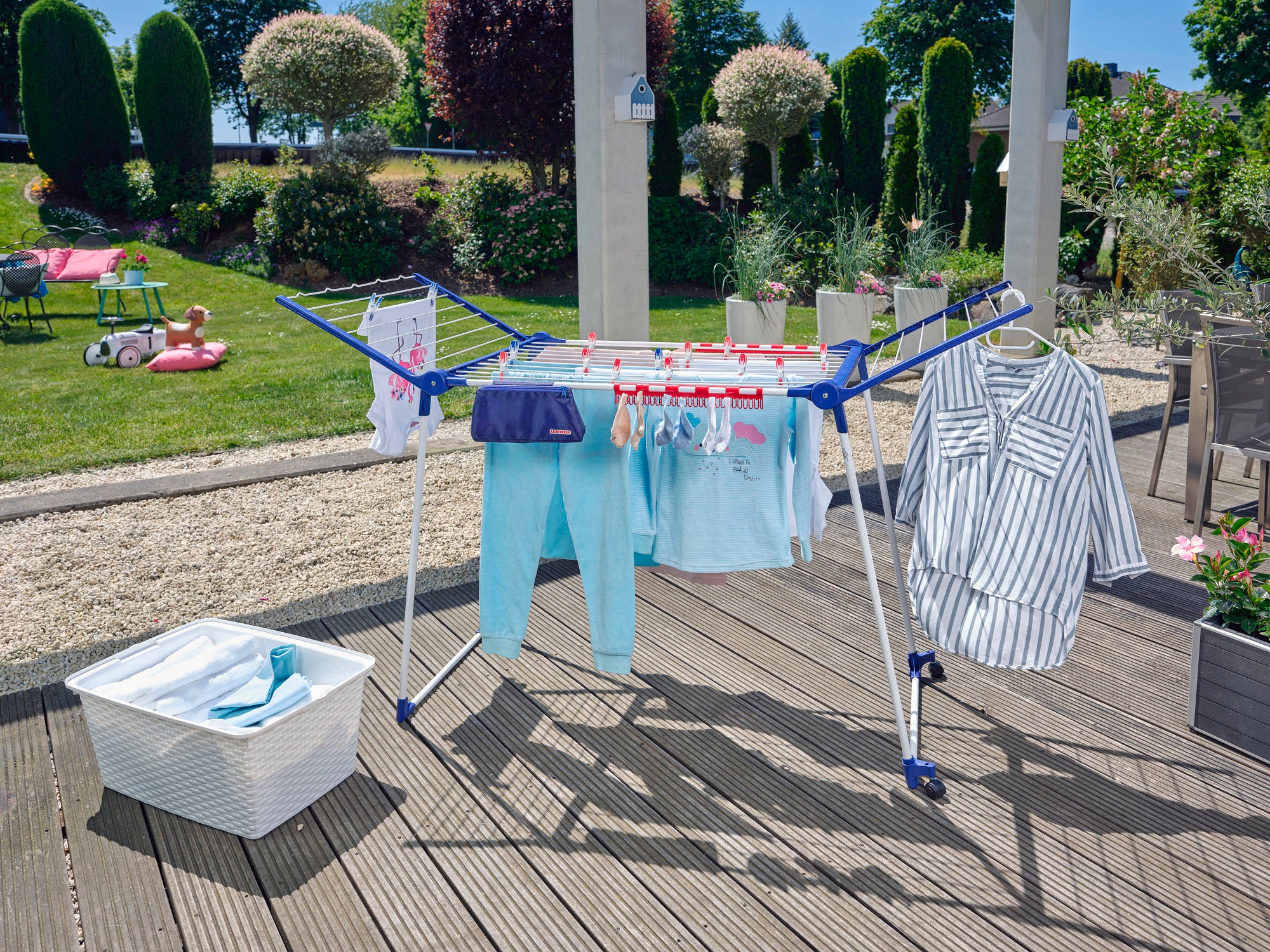 Standtrockner Pegasus 200 Solid Deluxe Mobile auf einer Terrasse mit aufgehängter Kleidung. Im Hintergrund ein Garten mit Bäumen und Sträuchern. Ideal für große Wäschemengen.