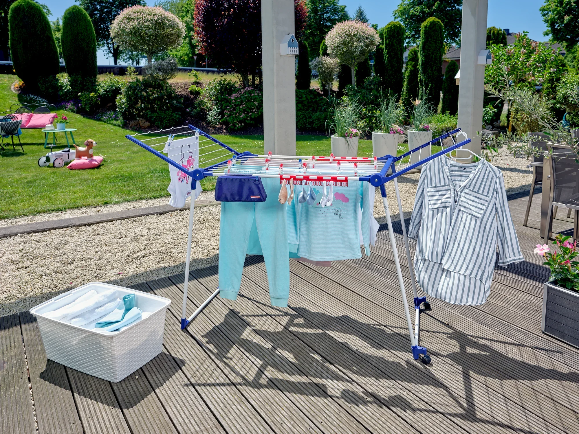 Suszarka Pegasus 200 Solid Deluxe Mobile na tarasie z wiszącymi ubraniami. W tle ogród z drzewami i krzewami. Idealna do dużych ilości prania.