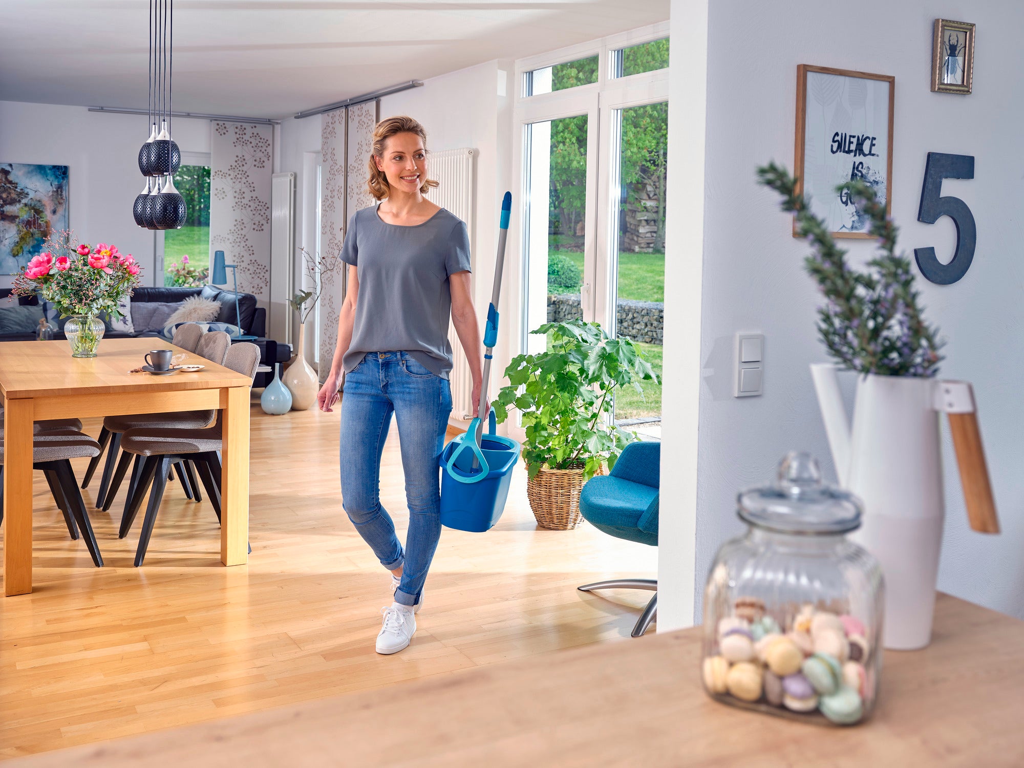 Eine Frau geht durch ein modernes Wohnzimmer und trägt das CLEAN TWIST Mop-Set. Der Raum ist hell und einladend, mit großen Fenstern und stilvoller Einrichtung. Pflanzen und Dekorationen verleihen dem Raum eine gemütliche Atmosphäre.