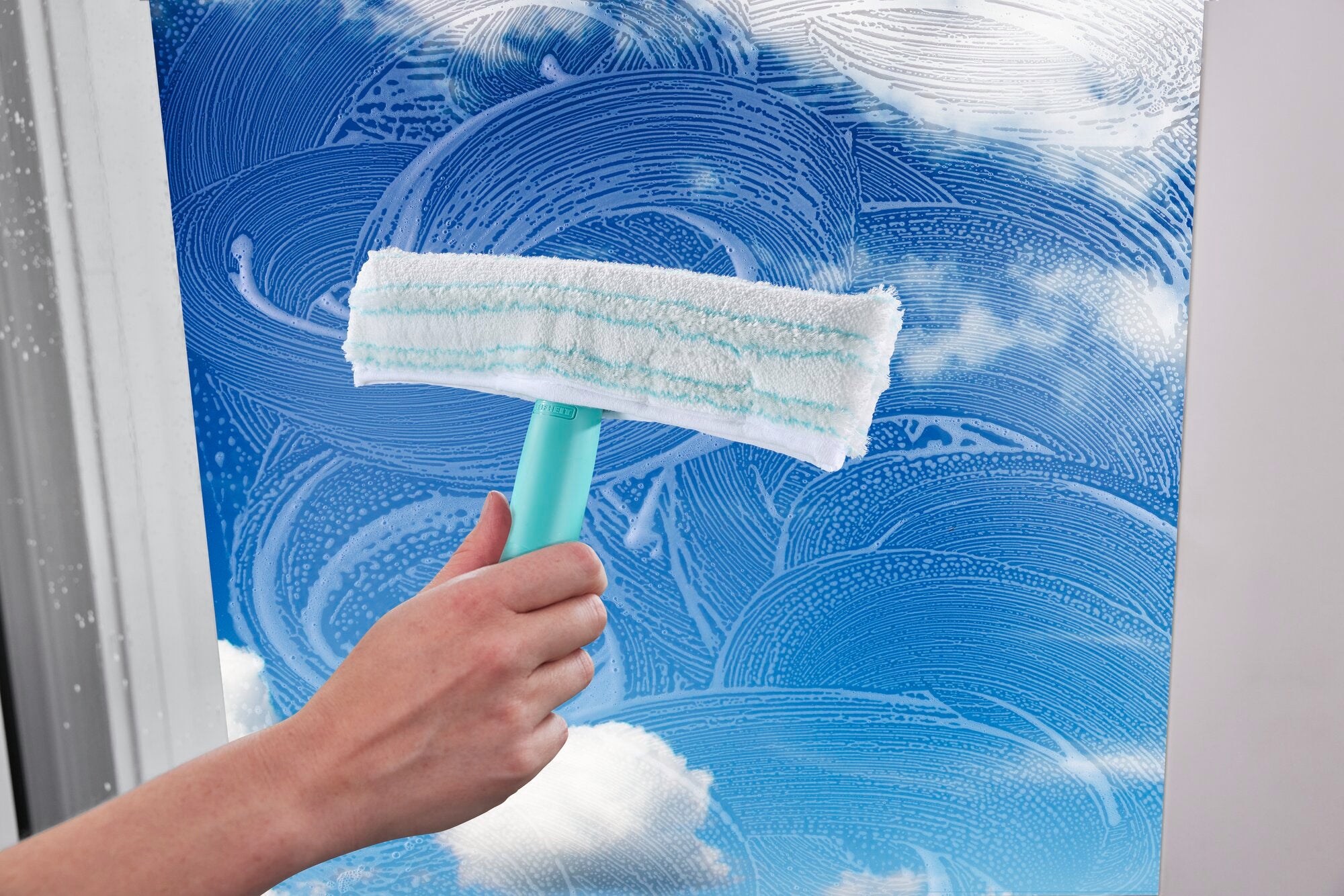 Een ruitensproeier wordt gebruikt om een raam schoon te maken. De hand houdt de sproeier vast terwijl het raam met ronddraaiende bewegingen van vuil wordt ontdaan.