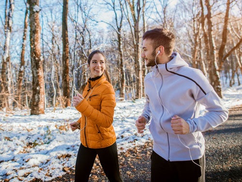Ein Mann und eine Frau joggen laechelnd auf einem schneebedeckten Waldweg; sie traegt eine orange Jacke, er eine graue.