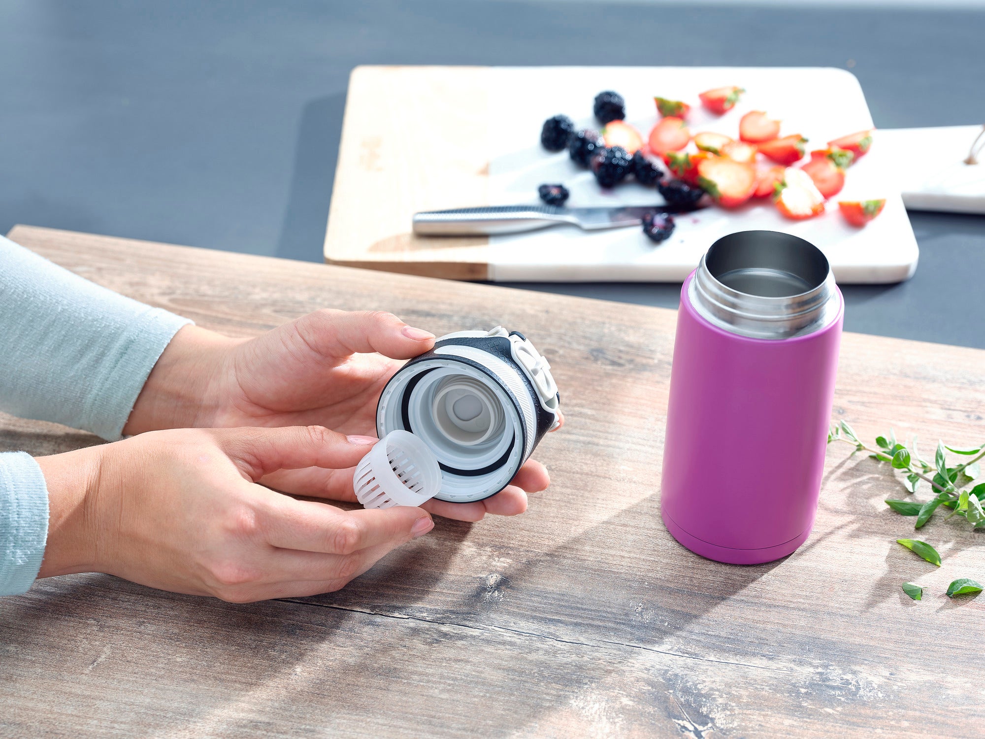 Isolierbecher Flip 350ml in Lila auf einem Tisch. Eine Person hält den Deckel des Bechers, während im Hintergrund ein Schneidebrett mit Obst zu sehen ist. Ideal für unterwegs.