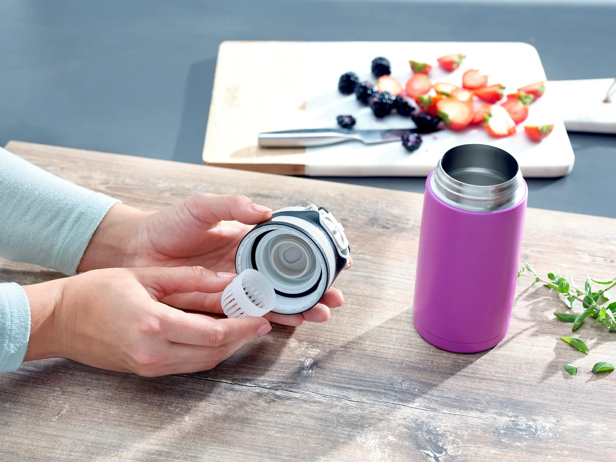 Fioletowy kubek próżniowy Flip 350 ml na stole. Osoba trzyma pokrywkę kubka, a w tle widać deskę do krojenia z owocami. Idealny w podróży.