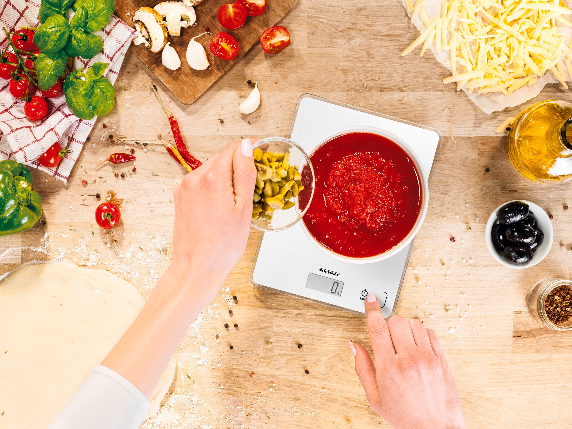 Eine digitale Küchenwaage zeigt 0 Gramm an, während eine Person gehackte Oliven in eine Schüssel mit Tomatensauce gibt. Um die Waage herum liegen Zutaten wie Käse, Olivenöl und Gemüse.