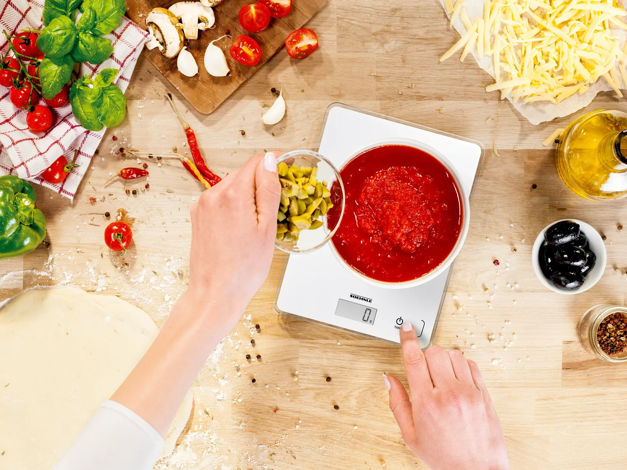 A digital kitchen scale displays 0 grams while a person adds chopped olives to a bowl of tomato sauce. Ingredients such as cheese, olive oil and vegetables are scattered around the scale.