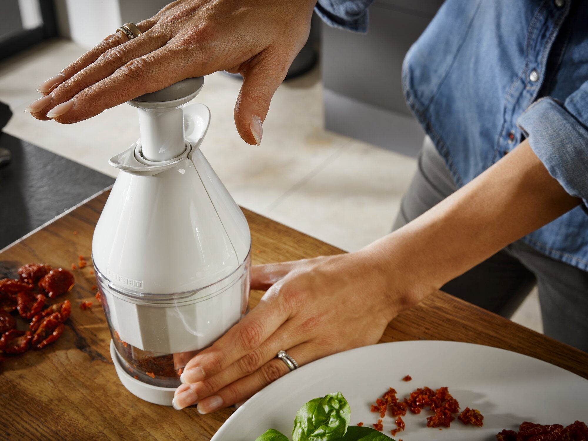 A person uses the onion and vegetable chopper to chop dried tomatoes on a wooden board. The chopper is white and is operated with one hand.