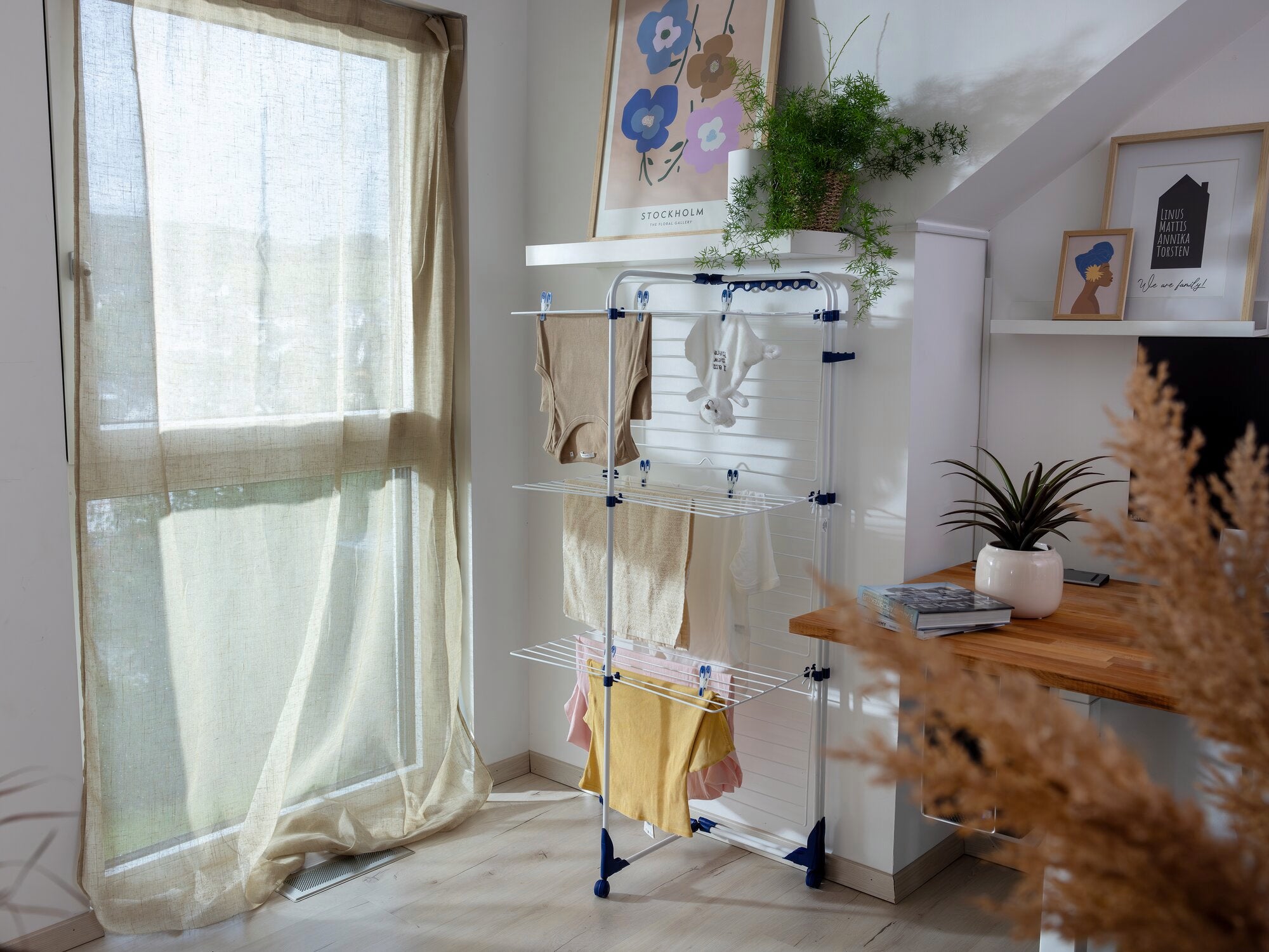 Een torendroger, de Classic Tower 340, staat in een lichte kamer naast een raam. Hij is volgeladen met wasgoed en omringd door planten en decoraties, wat een gezellige sfeer creëert.
