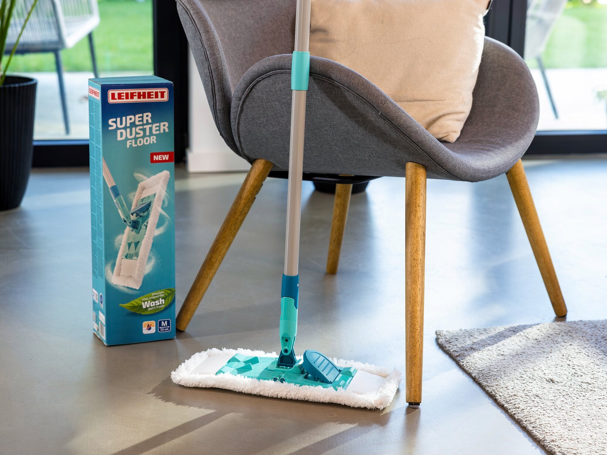 Ein moderner Staubwischer, der SUPERDUSTER Floor, steht neben einer Verpackung in einem stilvollen Raum. Der Wischer hat einen langen Stiel und ein breites, flaches Wischtuch. Ideal für die Reinigung großer Flächen.