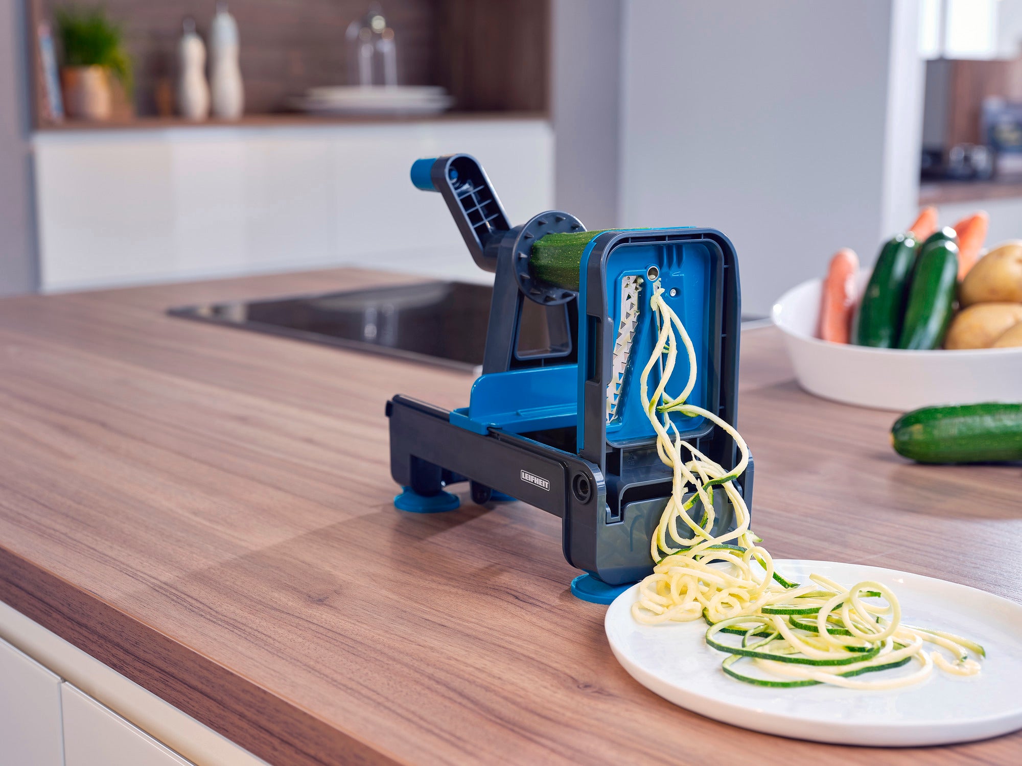 A blue tabletop spiral slicer on a kitchen worktop cuts courgettes into spiral-shaped vegetable strips. More fresh ingredients can be seen in the background.