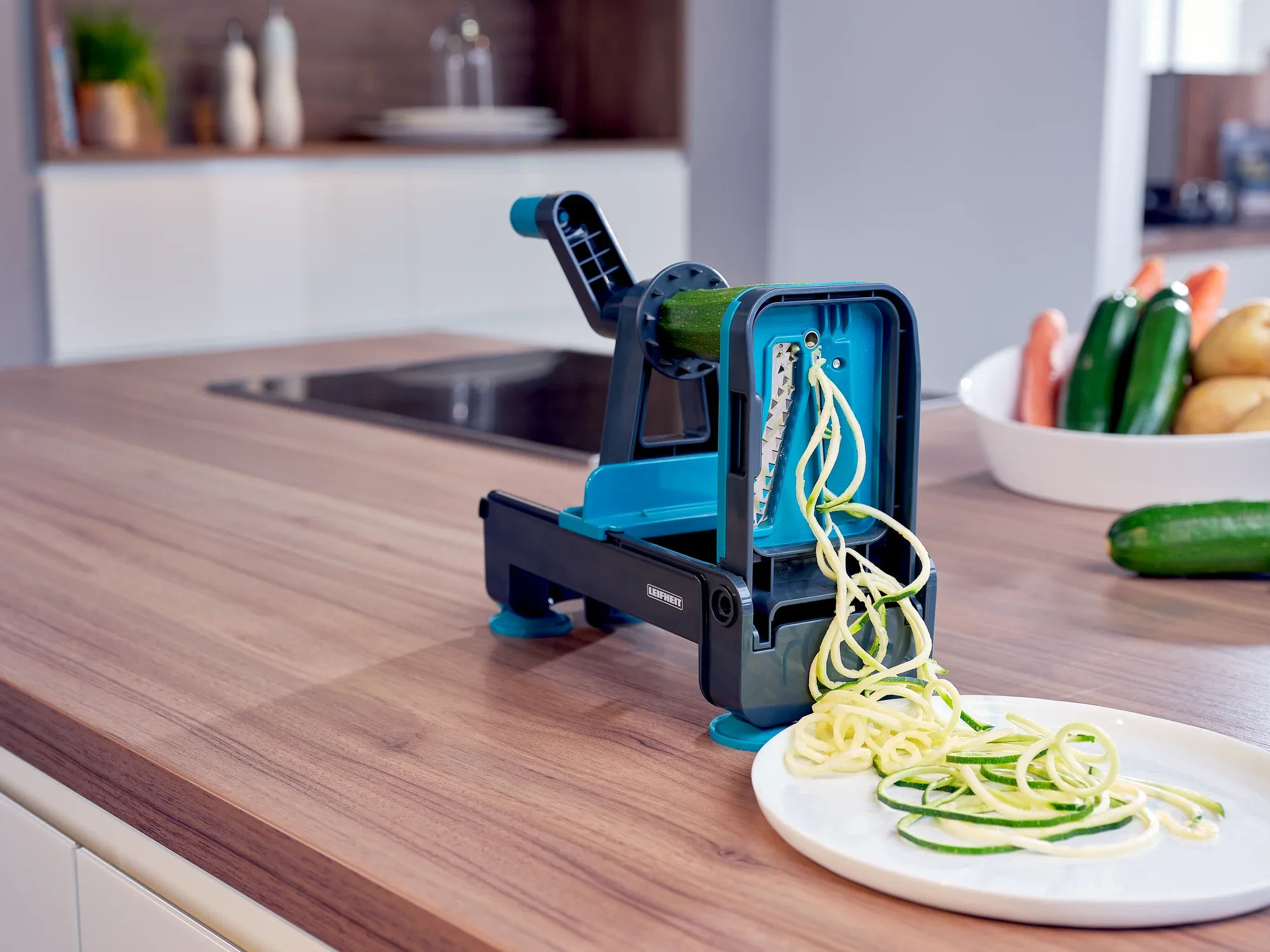 Ein blauer Tisch-Spiralschneider auf einer Küchenarbeitsplatte schneidet Zucchini in spiralförmige Gemüsestreifen. Im Hintergrund sind weitere frische Zutaten zu sehen.