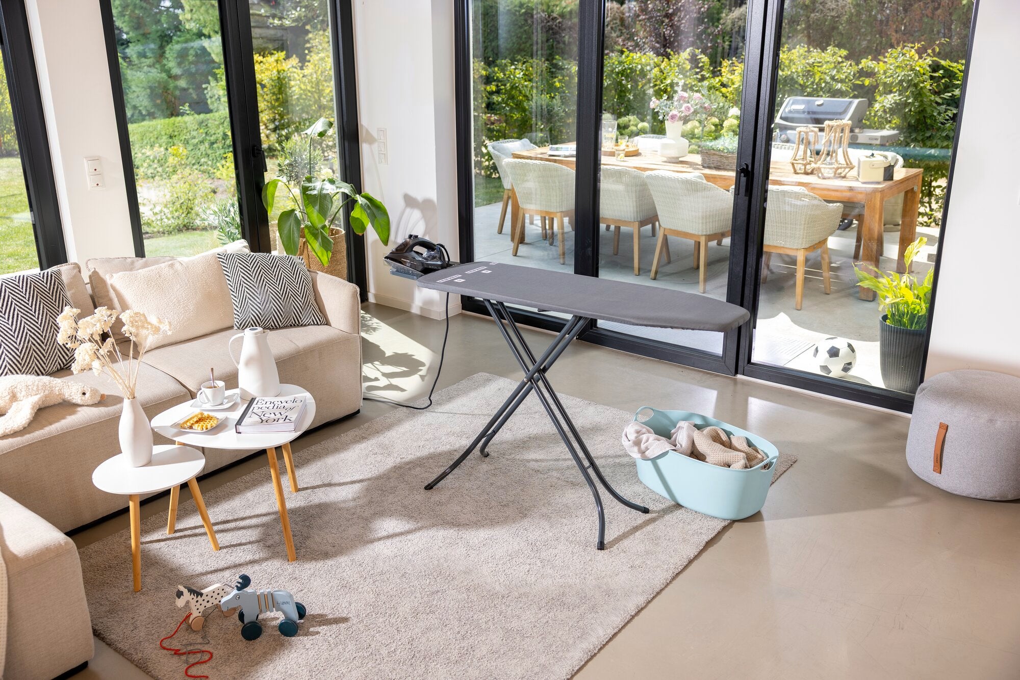 Bügelbrett Leifheit Classic M in Schwarz steht in einem modernen Wohnzimmer mit großen Fenstern. Im Hintergrund ist ein Garten zu sehen. Auf dem Tisch liegen Dekorationsartikel.