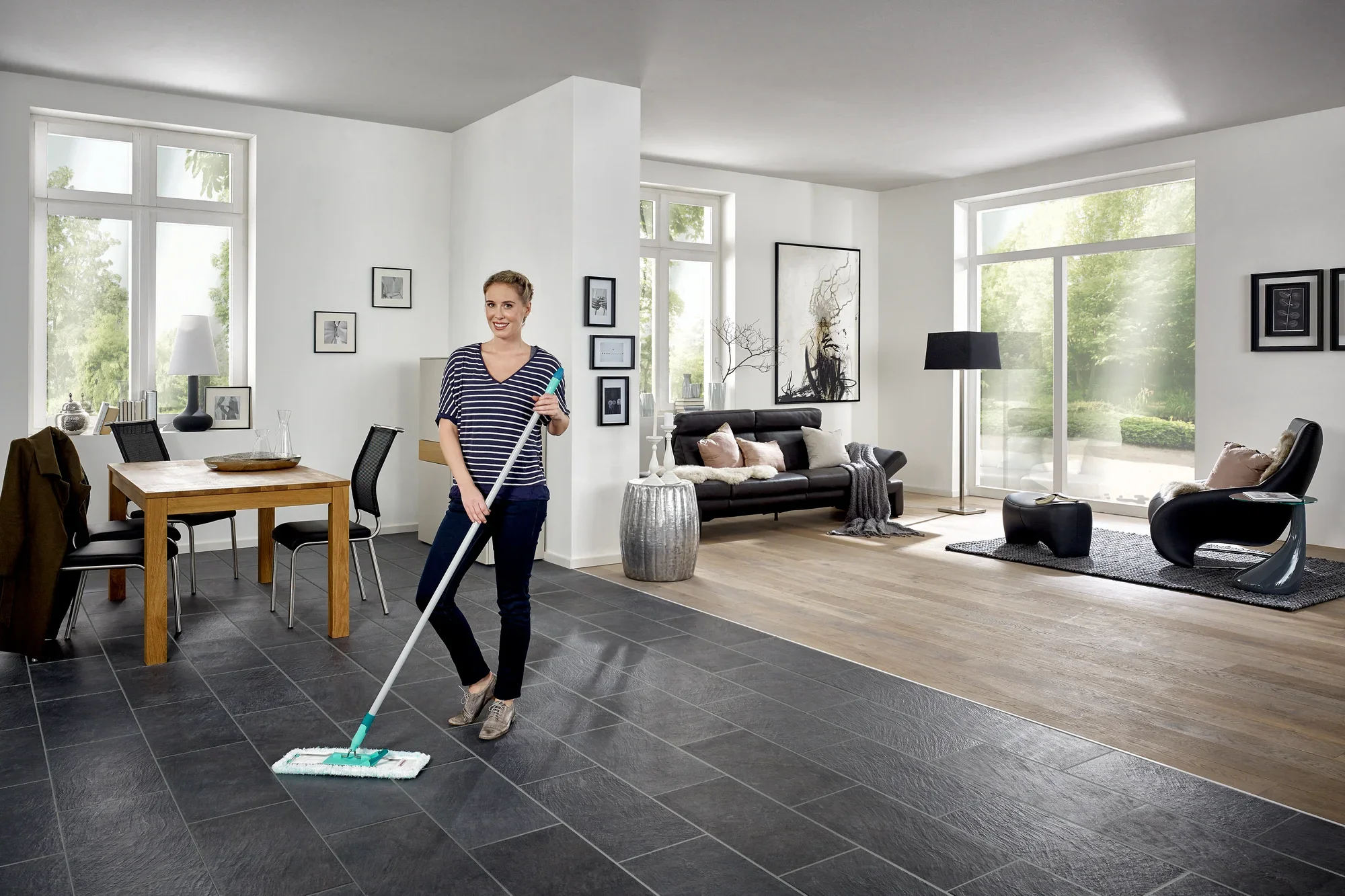 Una donna pulisce il pavimento di un moderno soggiorno con il lavapavimenti Profi XL. La stanza è luminosa e arredata con stile, con grandi finestre e mobili eleganti.