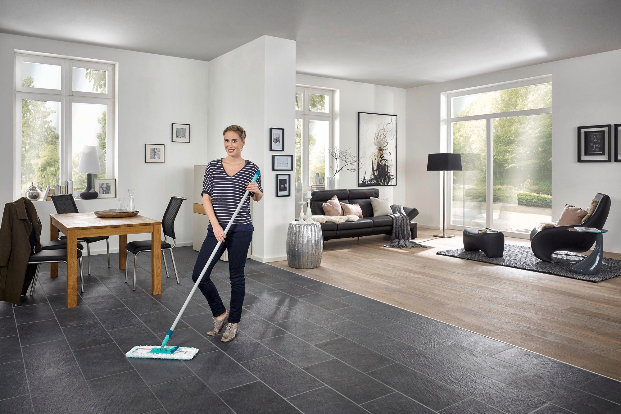 A woman is mopping the floor of a modern living room with the Profi XL floor mop. The room is bright and stylishly furnished, with large windows and elegant furniture.