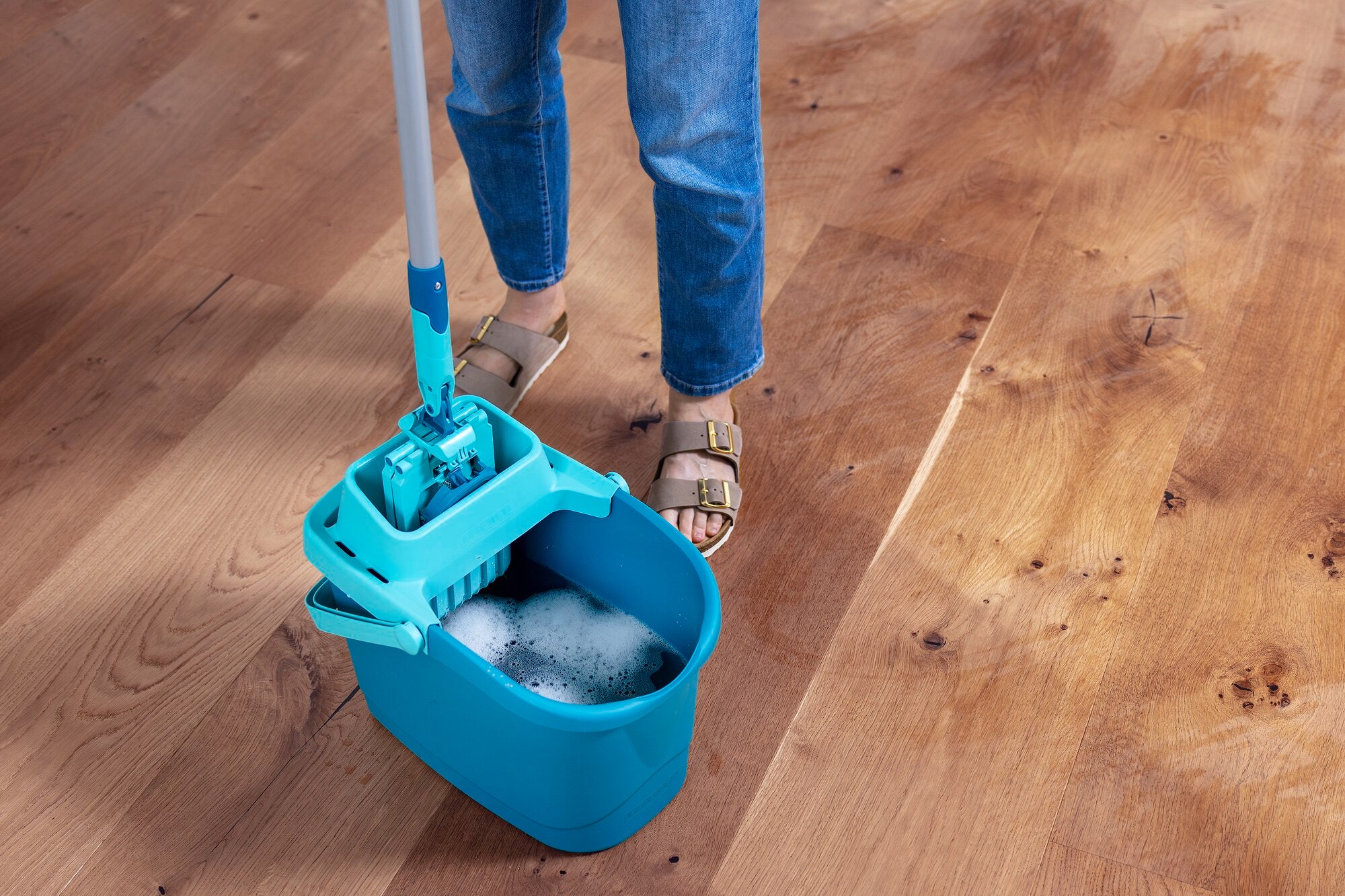 Una persona limpia un suelo de madera con el set POWER CLEAN. El cubo está lleno de agua jabonosa y la mopa se sumerge en él. La escena muestra una limpieza eficaz del suelo.