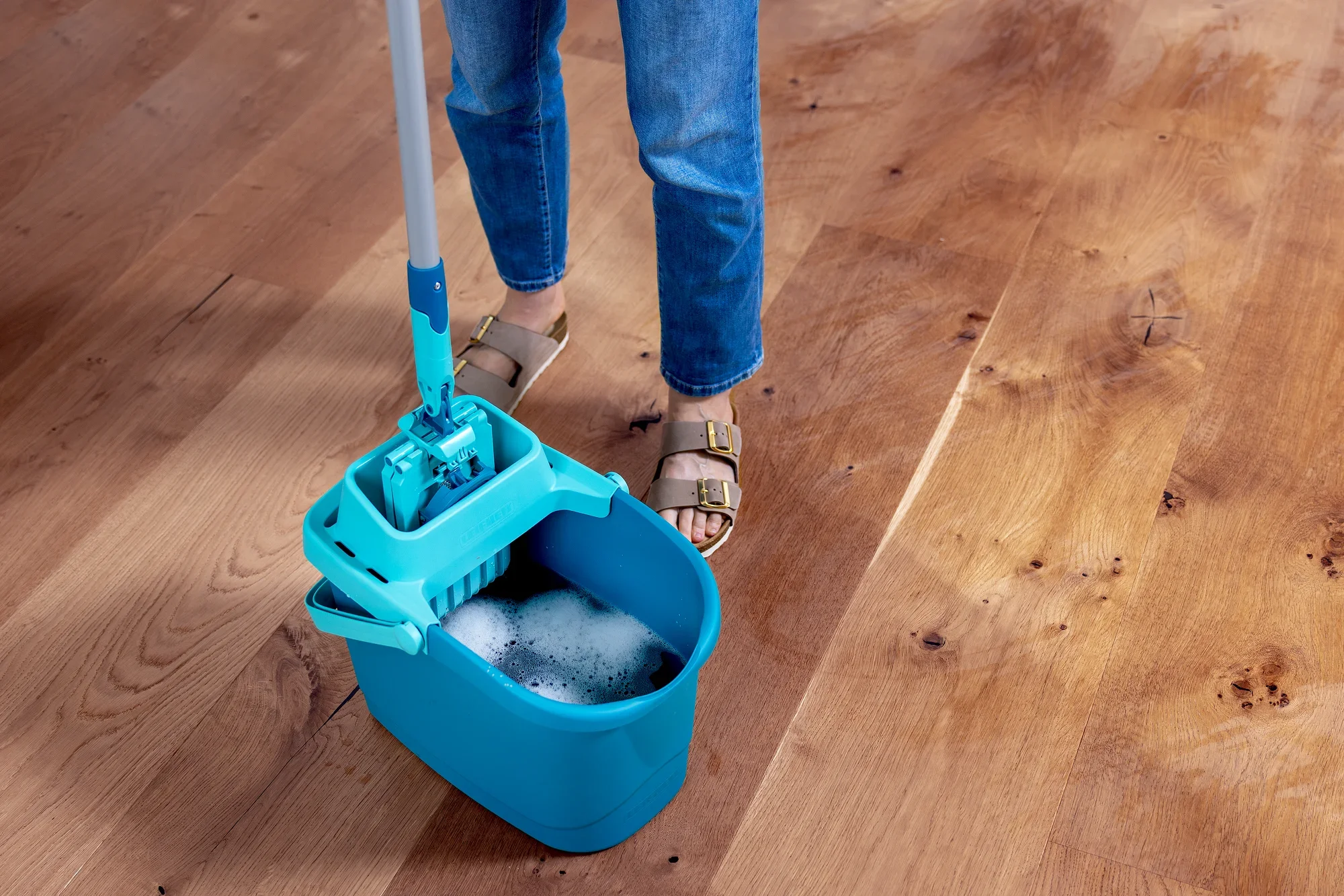 Une personne nettoie un sol en bois avec le kit POWER CLEAN. Le seau est rempli d'eau savonneuse et la serpillière est plongée dans le seau. La scène montre un nettoyage efficace du sol.