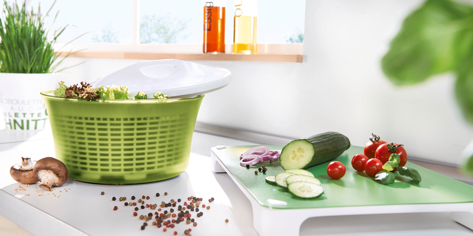 Salatschleuder 5,5l in einer modernen Küche. Die grüne Schleuder ist mit frischem Salat gefüllt. Daneben liegen geschnittene Gurken, Tomaten und Zwiebeln auf einem Schneidebrett.
