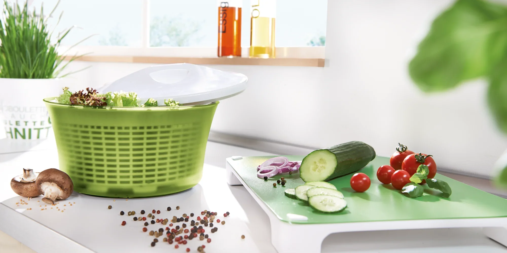 Salaterka o pojemności 5,5 litra w nowoczesnej kuchni. Zielony pojemnik jest wypełniony świeżą sałatą. Obok na desce do krojenia leżą pokrojone ogórki, pomidory i cebula.