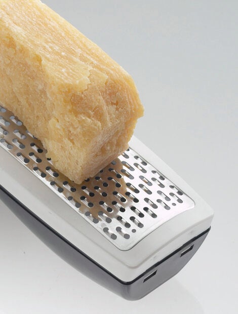 Eine Parmesanreibe wird verwendet, um ein großes Stück Parmesan zu reiben. Die Reibe hat ein modernes Design mit einem ergonomischen Griff und einer robusten Metallfläche.
