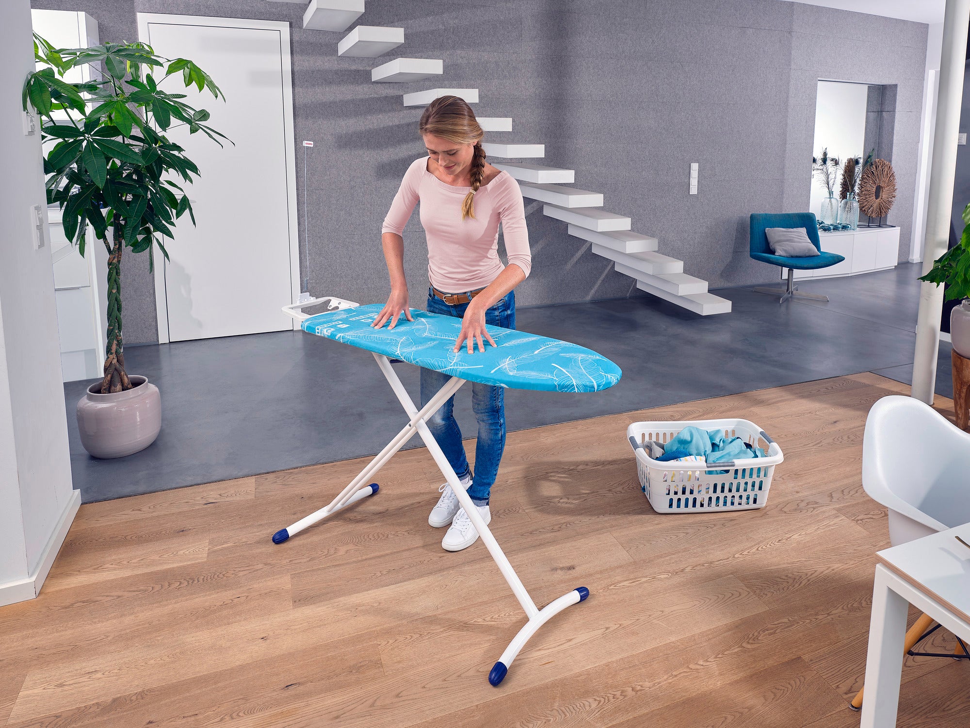 Eine Frau bügelt auf einem Leifheit Air Board M Solid Bügeltisch in einem modernen Wohnzimmer. Der Tisch ist blau und steht auf einem Holzboden. Im Hintergrund ist ein Korb mit Wäsche.