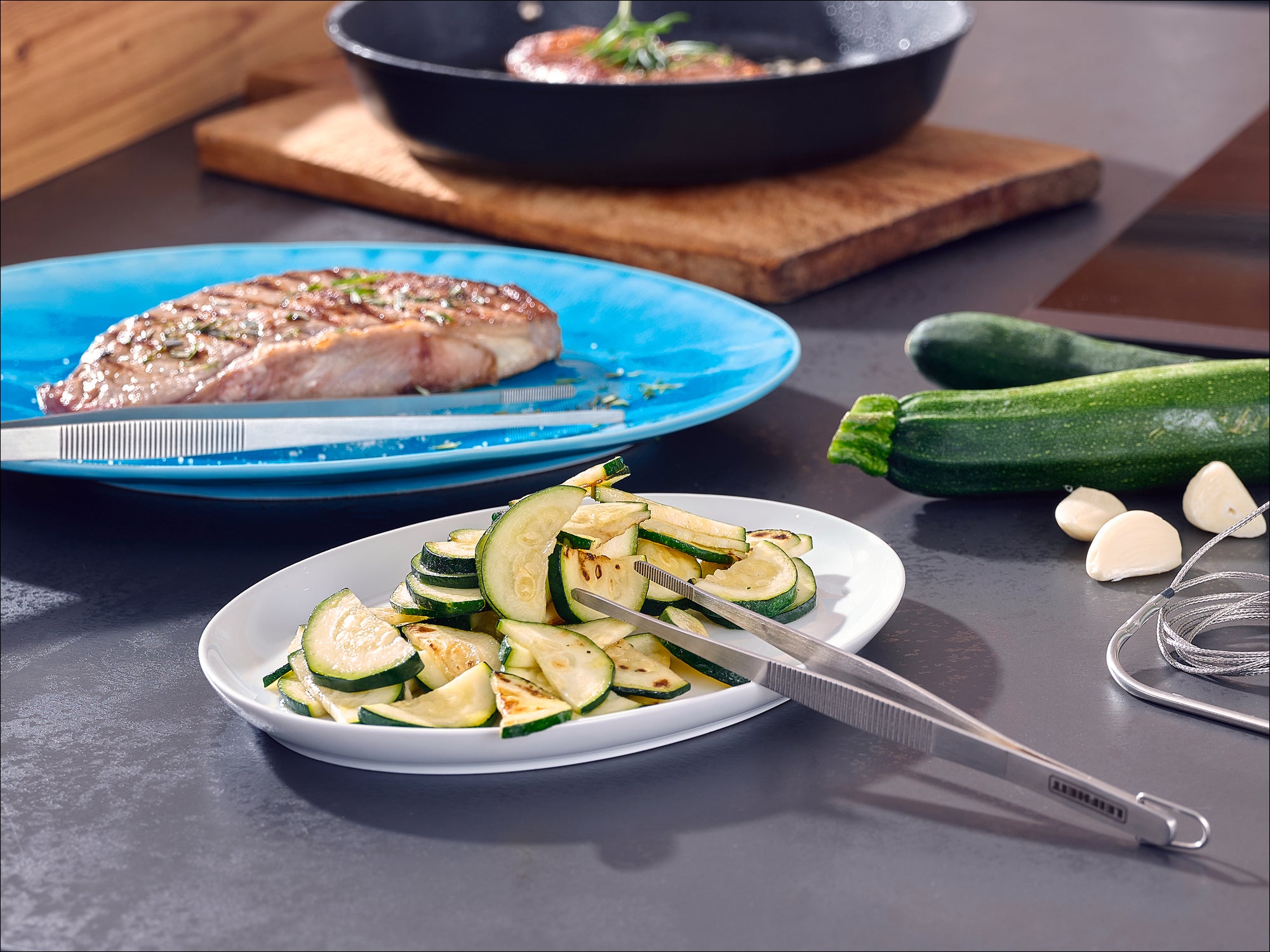 Auf einem Tisch liegen ein Teller mit gegrillten Zucchinischeiben und eine Grillzange. Im Hintergrund ist ein Teller mit einem Steak und eine Pfanne zu sehen. Frische Zucchini und Knoblauch liegen daneben.