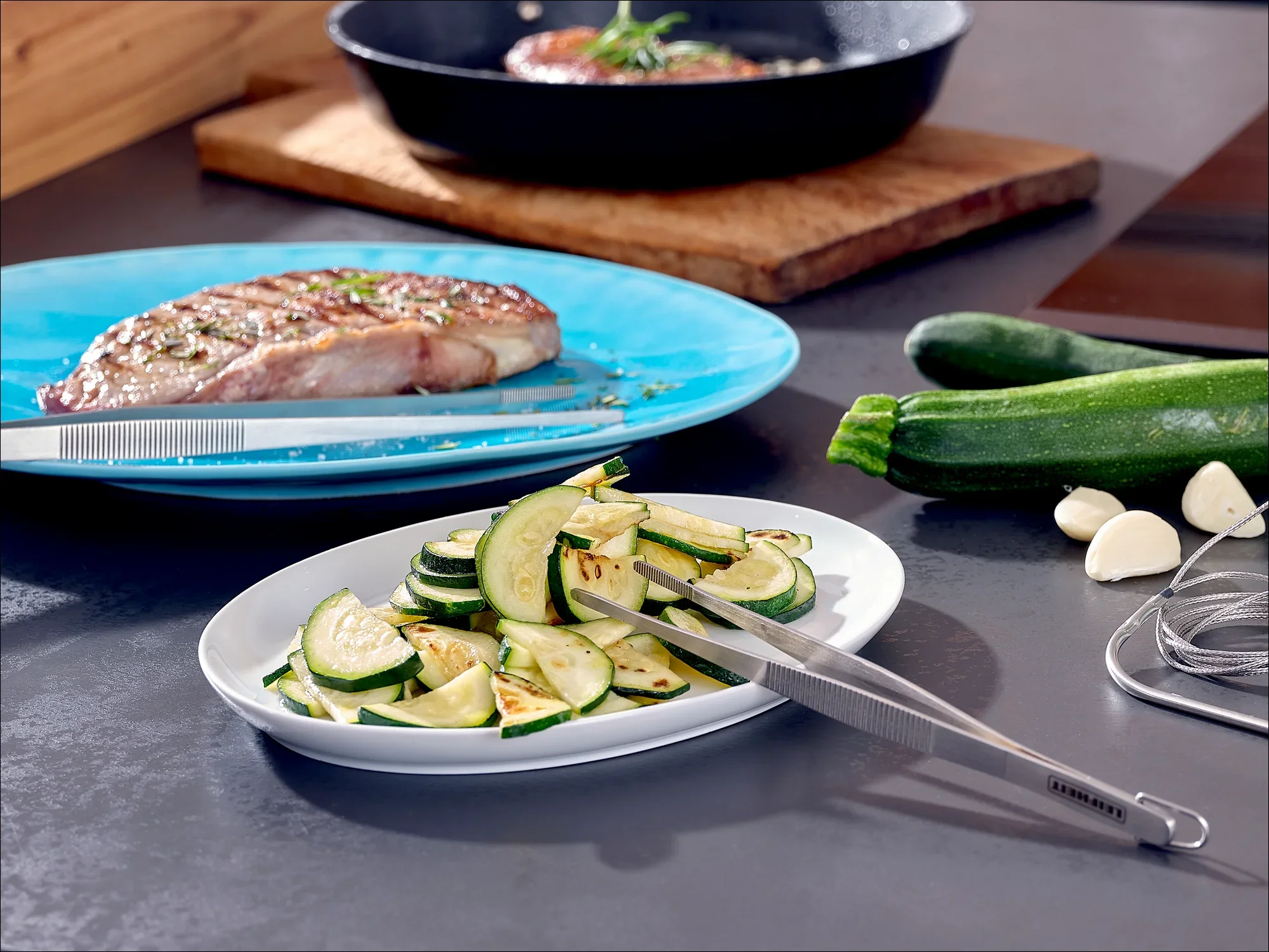 Op tafel staat een bord met gegrilde courgetteplakjes en een grilltang. Op de achtergrond is een bord met een biefstuk en een pan te zien. Verse courgettes en knoflook liggen ernaast.