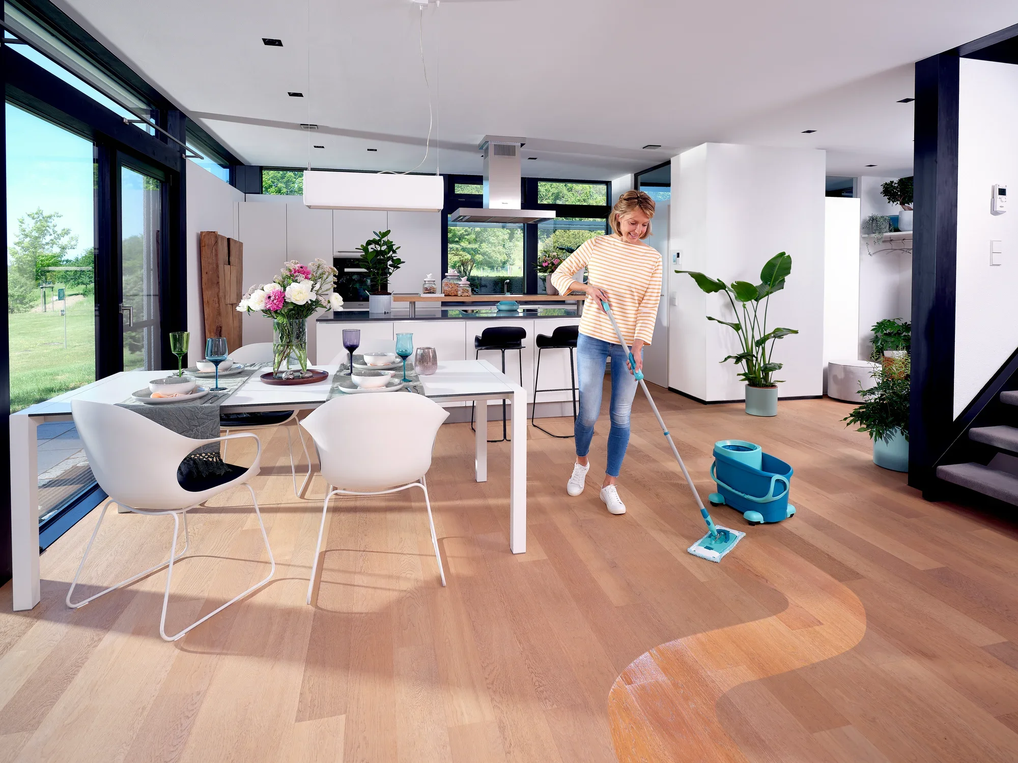 Een vrouw reinigt de houten vloer in een moderne keuken met de CLEAN TWIST M Ergo Set. De kamer is licht en stijlvol ingericht, met grote ramen en planten.