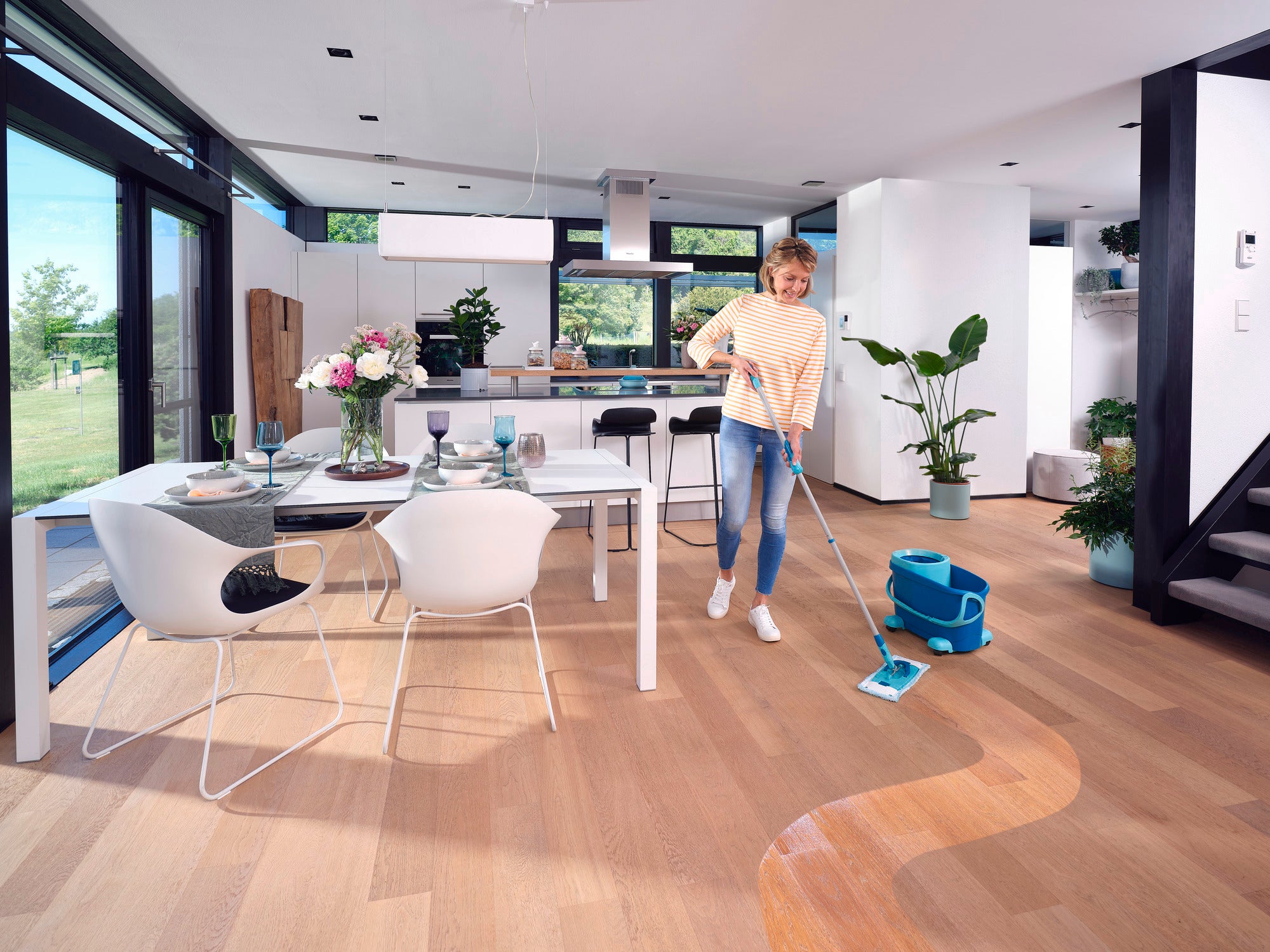 Een vrouw reinigt de houten vloer in een moderne keuken met de CLEAN TWIST M Ergo Set. De kamer is licht en stijlvol ingericht, met grote ramen en planten.