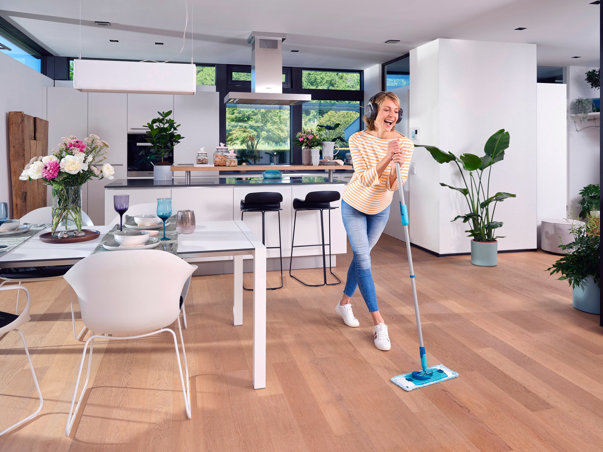 Een vrouw dweilt met de CLEAN TWIST M Ergo in een moderne, lichtgekleurde keuken. De vloer is van hout en planten en bloemen sieren de kamer. Ze ziet er vrolijk en ontspannen uit.