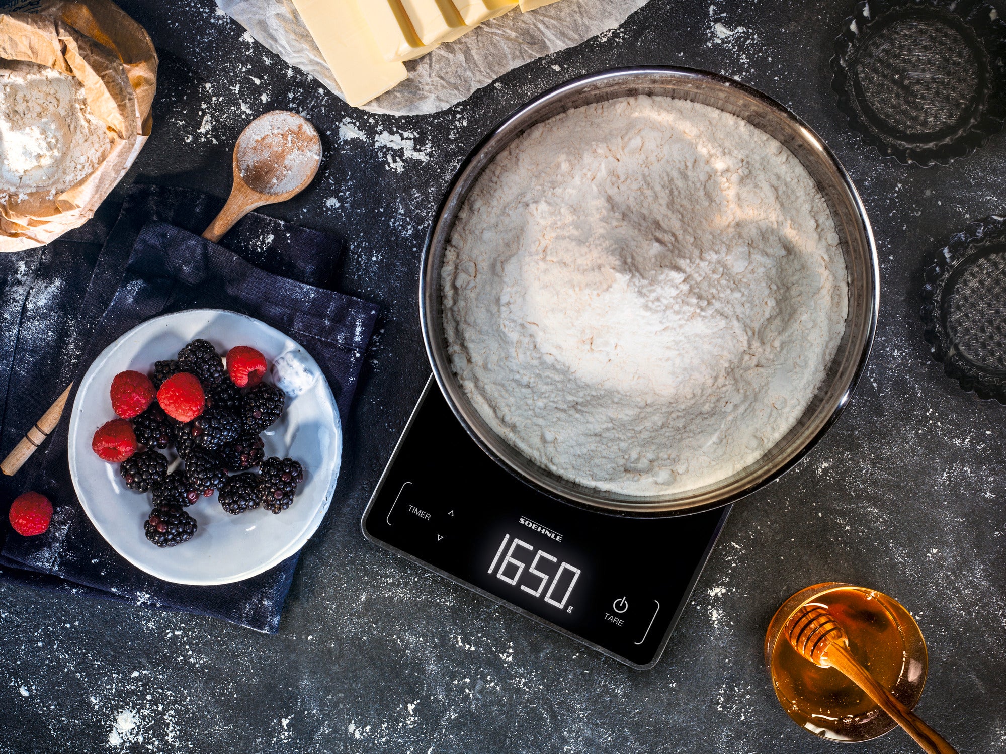 A digital kitchen scale shows 1650 grams of flour in a bowl. Next to it are fresh berries on a plate, a wooden spoon and a jar of honey on a dark work surface.