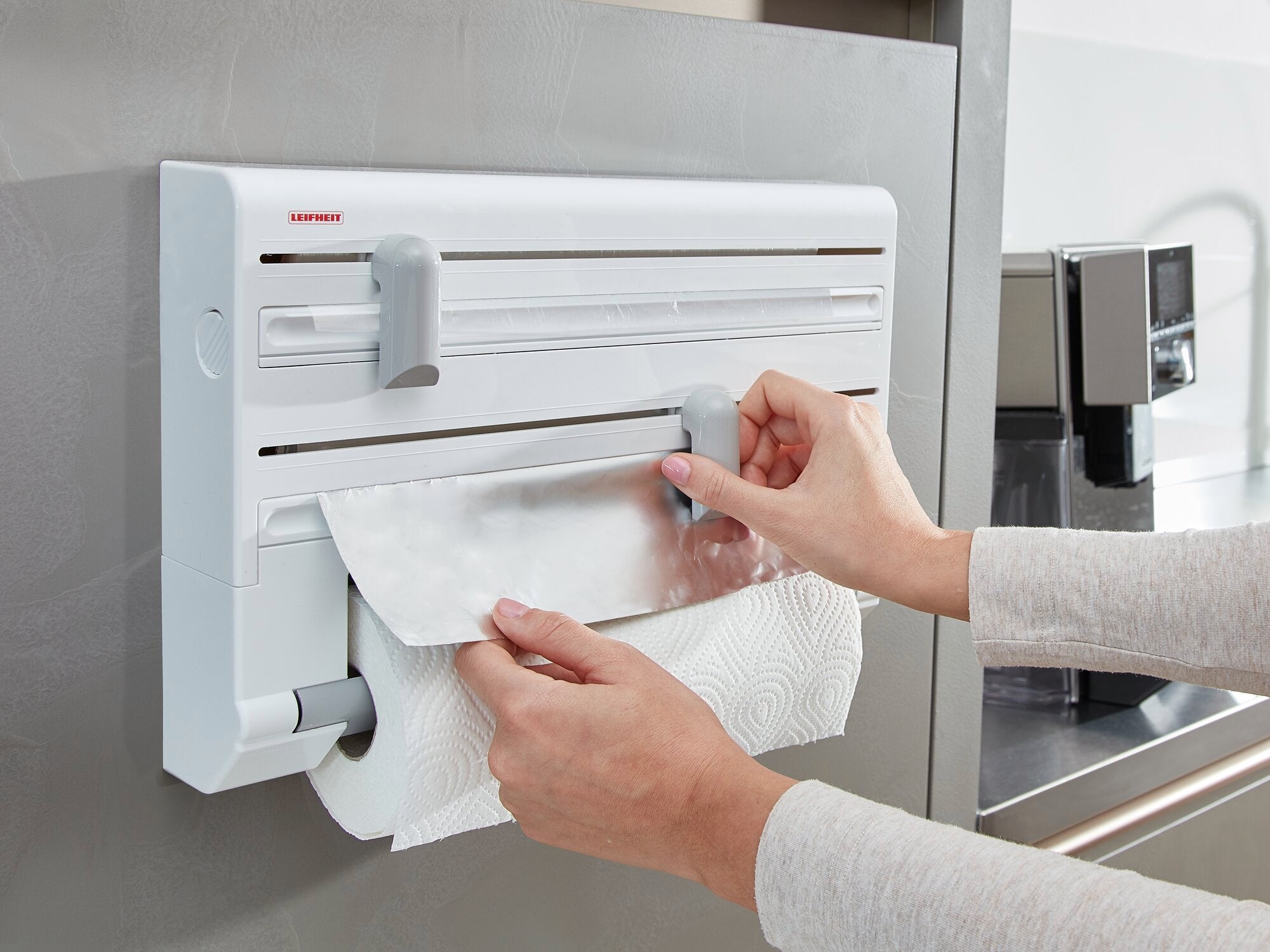 Parat wall-mounted roll holder from Leifheit mounted on a kitchen wall. A person pulls aluminium foil from the holder while a kitchen roll hangs underneath. Ideal for efficient kitchen organisation.