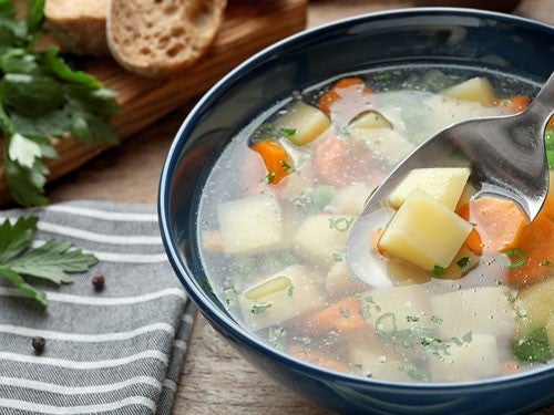 Eine Schuessel Kartoffelsuppe mit Kartoffel- und Karottenstuecken, garniert mit frischen Kraeutern, daneben Brot und Petersilie auf einem Holzbrett.