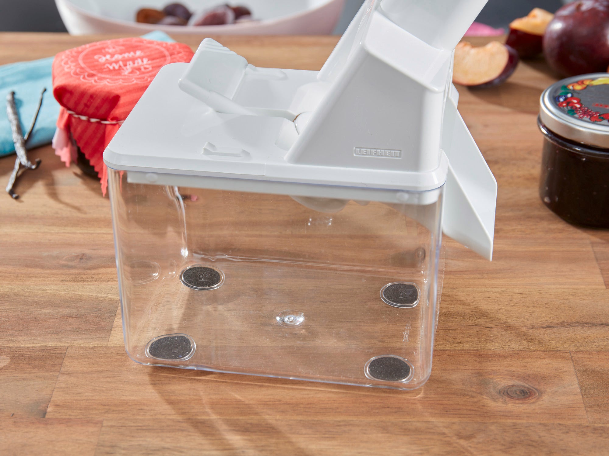 Uno snocciolatore di prugne su un tavolo di legno. Il dispositivo ha un coperchio bianco e un contenitore trasparente. Sullo sfondo si vedono prugne e marmellata. Ideale per snocciolare facilmente le prugne.