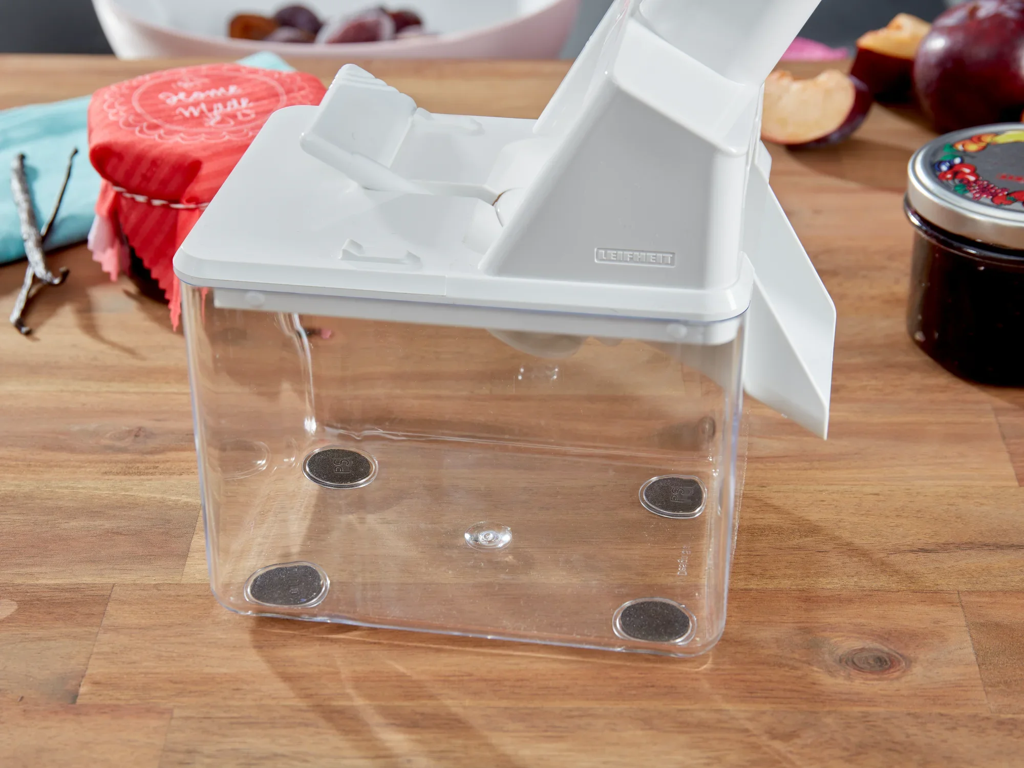 Destonator do śliwek na drewnianym stole. Urządzenie ma białą pokrywę i przezroczysty pojemnik. W tle widać śliwki i dżem. Idealny do łatwego drylowania śliwek.