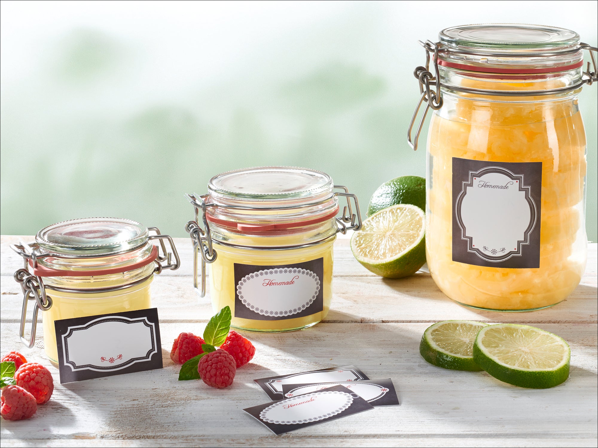 Drei Einmachgläser mit Etiketten auf einem Holztisch, umgeben von frischen Limetten und Himbeeren. Die Gläser enthalten eine gelbe Substanz, möglicherweise Marmelade oder Gelee.