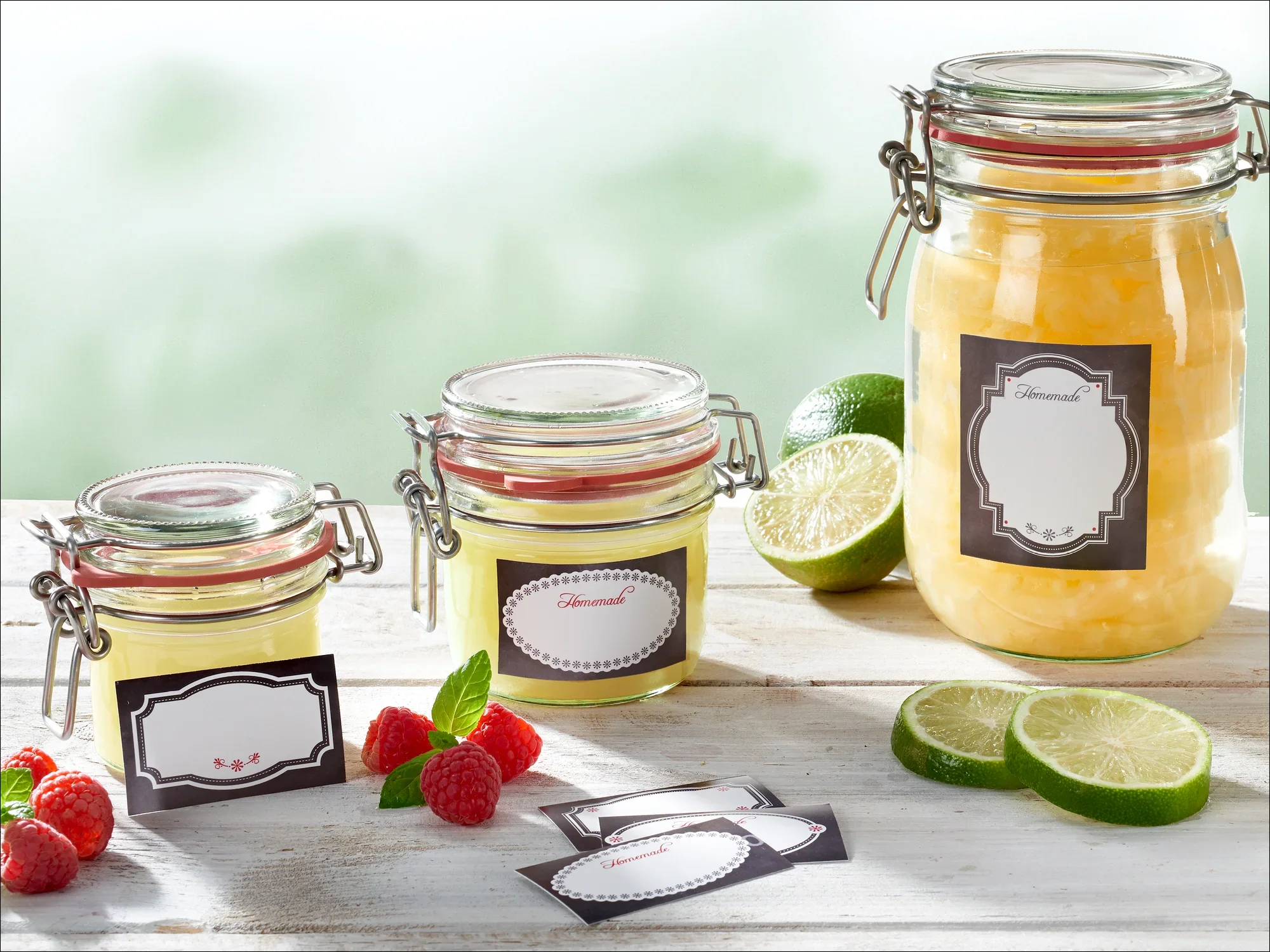 Drie inmaakpotten met etiketten op een houten tafel, omringd door verse limoenen en frambozen. De potten bevatten een gele substantie, mogelijk jam of gelei.