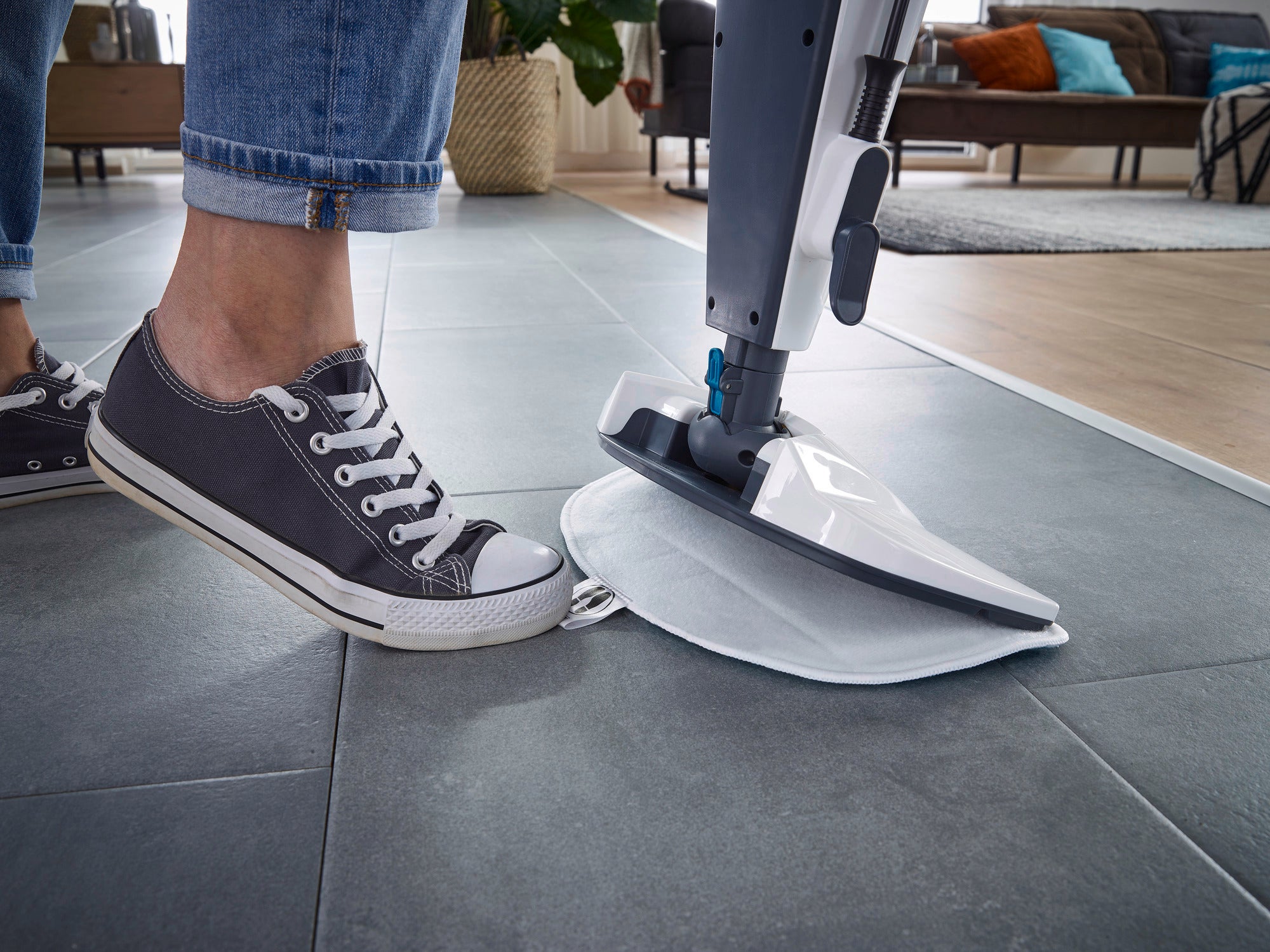 Dampfreiniger CleanTenso Power in Nahaufnahme auf einem grauen Fliesenboden. Eine Person mit Sneakers bedient den Reiniger, während im Hintergrund ein gemütliches Wohnzimmer zu sehen ist.