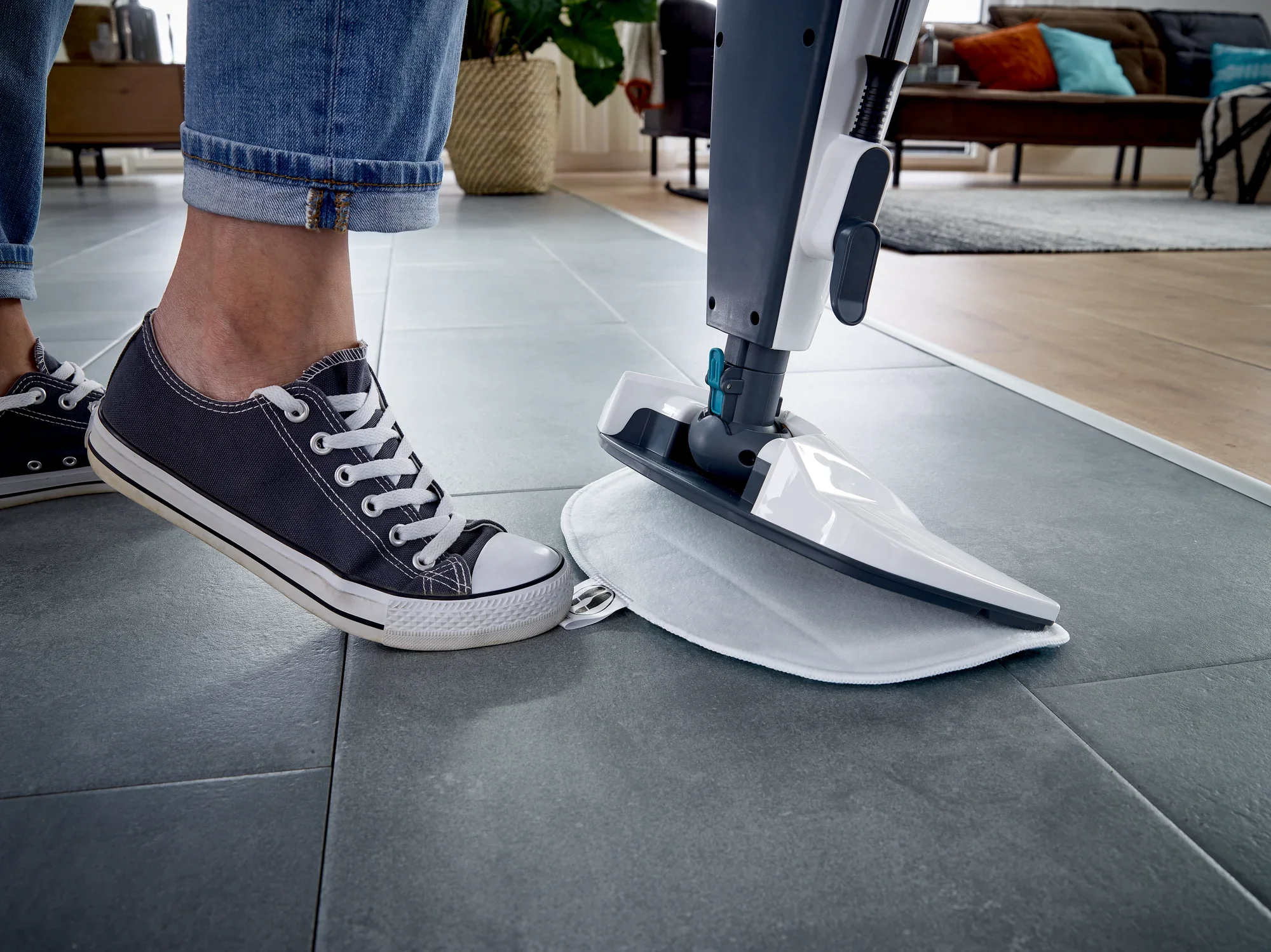 Gros plan sur le nettoyeur vapeur CleanTenso Power posé sur un sol carrelé gris. Une personne en baskets utilise le nettoyeur, tandis qu'un salon confortable est visible en arrière-plan.