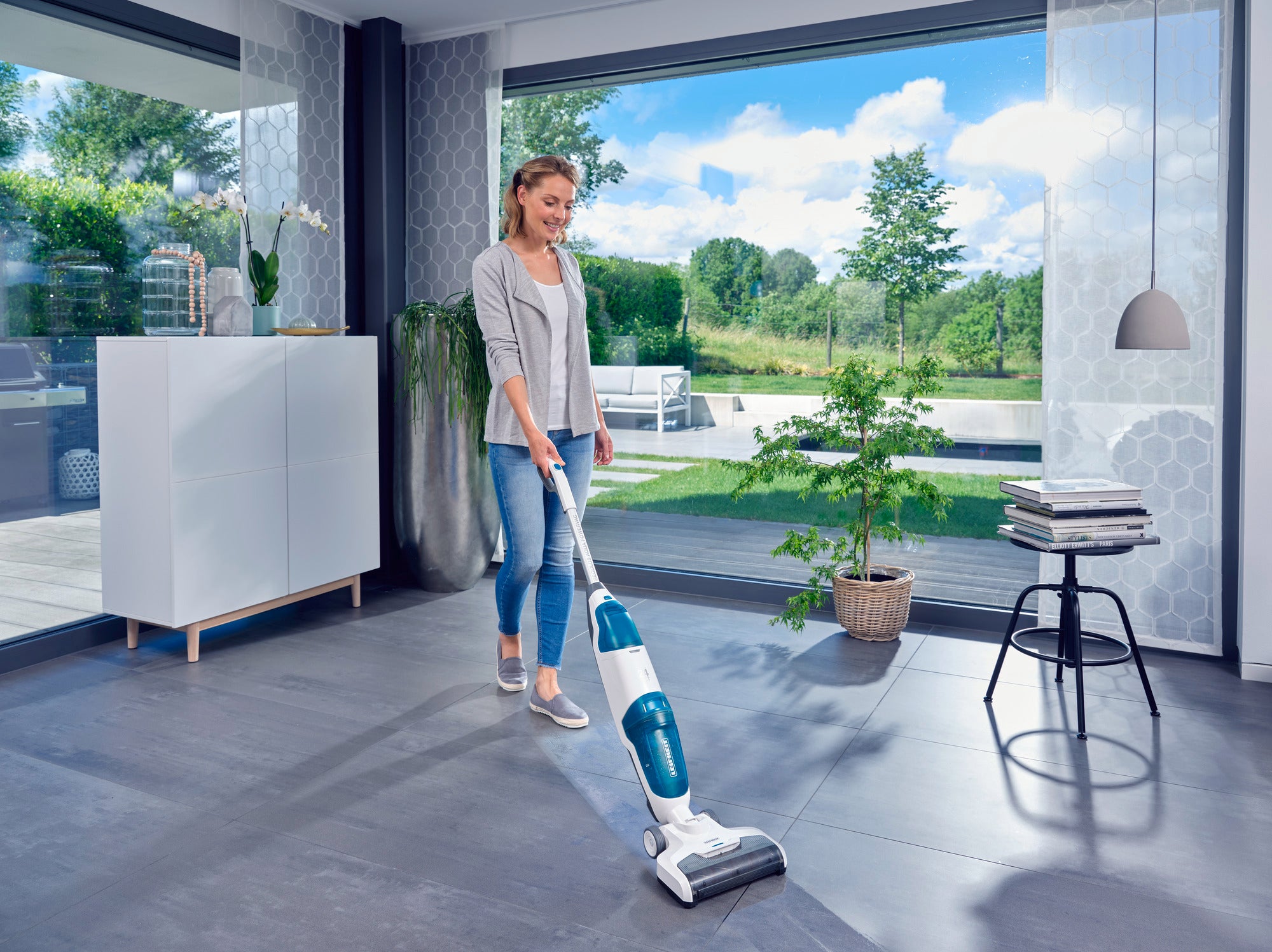 Eine Frau reinigt den Boden eines modernen Wohnzimmers mit dem Regulus Aqua PowerVac. Große Fenster bieten einen Blick auf einen sonnigen Garten. Der Raum wirkt hell und einladend.