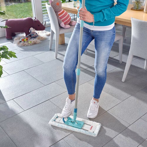 Eine Person reinigt einen gefliesten Boden mit einem Leifheit Bodenwischer. Im Hintergrund sind ein Tisch, Stühle und ein Spielzeug zu sehen. Die Szene wirkt hell und einladend.