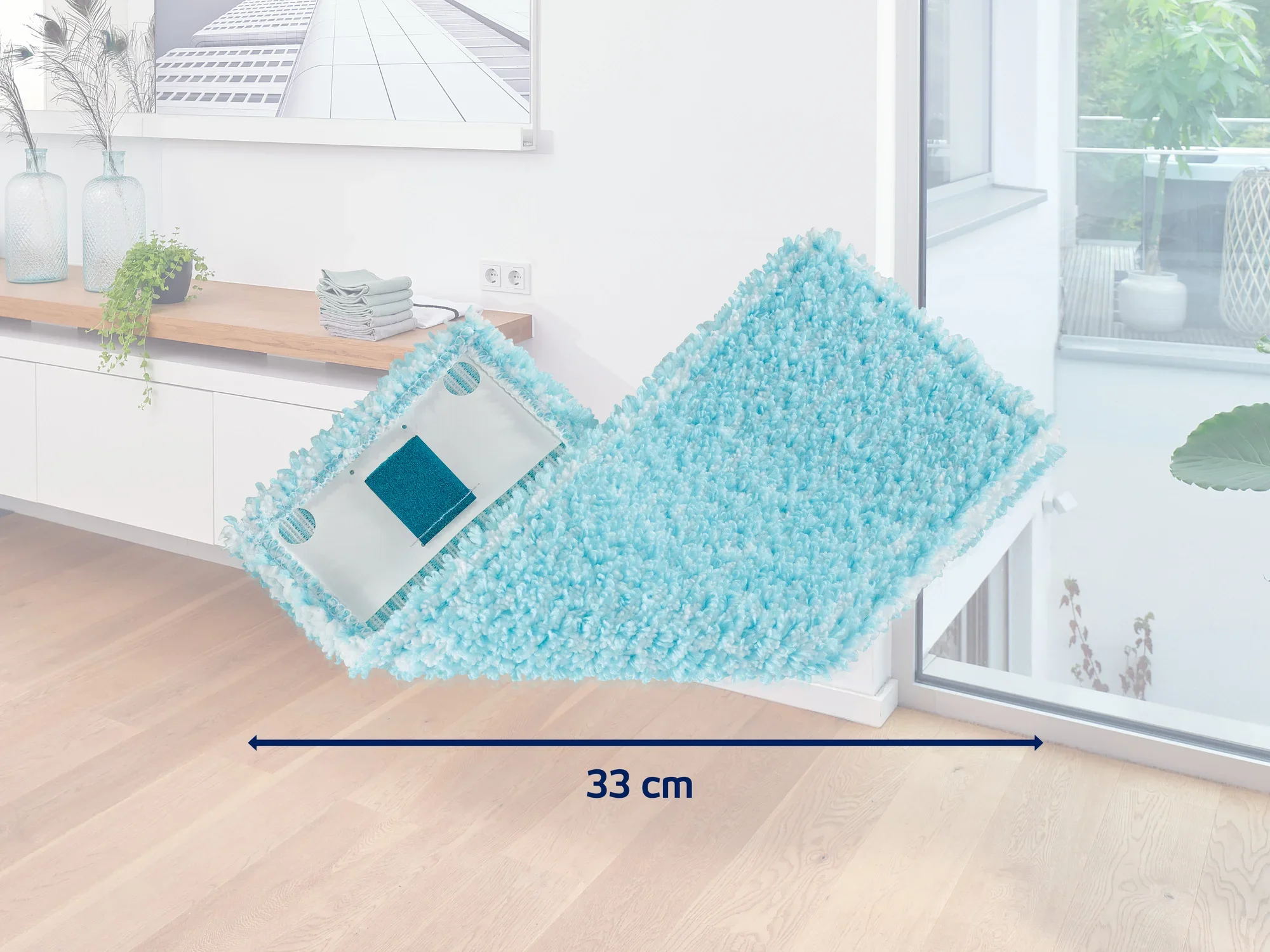 Een turquoise gekleurde mop met een breedte van 33 cm ligt op een lichtgekleurde houten vloer in een moderne kamer. Op de achtergrond zijn een dressoir en grote ramen te zien.