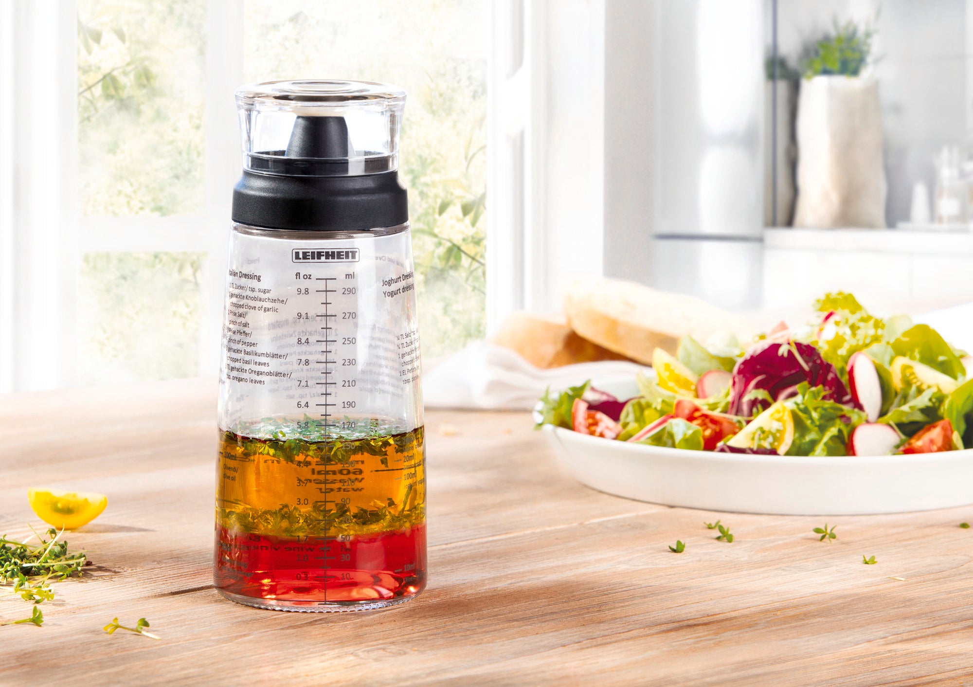 Ein Salat-Dressing-Shaker steht auf einem Holztisch in einer hellen Küche. Im Hintergrund ist ein frischer Salat zu sehen. Der Shaker hat Messskalen und einen schwarzen Deckel.