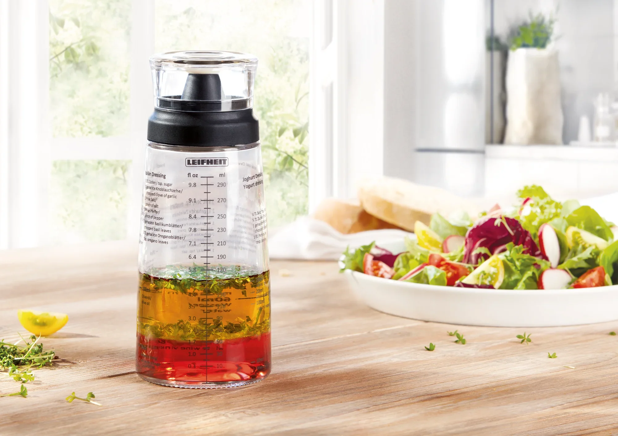 Een saladedressing shaker staat op een houten tafel in een lichtgekleurde keuken. Op de achtergrond is een verse salade te zien. De shaker heeft een maatverdeling en een zwart deksel.
