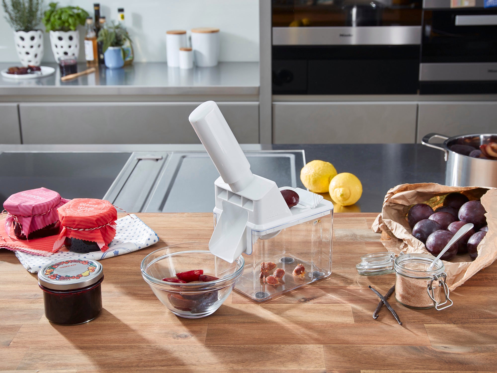 Un dénoyauteur de prunes posé sur un plan de travail moderne. Entouré de prunes fraîches, de pots de confiture et d'ustensiles de cuisine, l'image montre une scène pratique dans une cuisine.