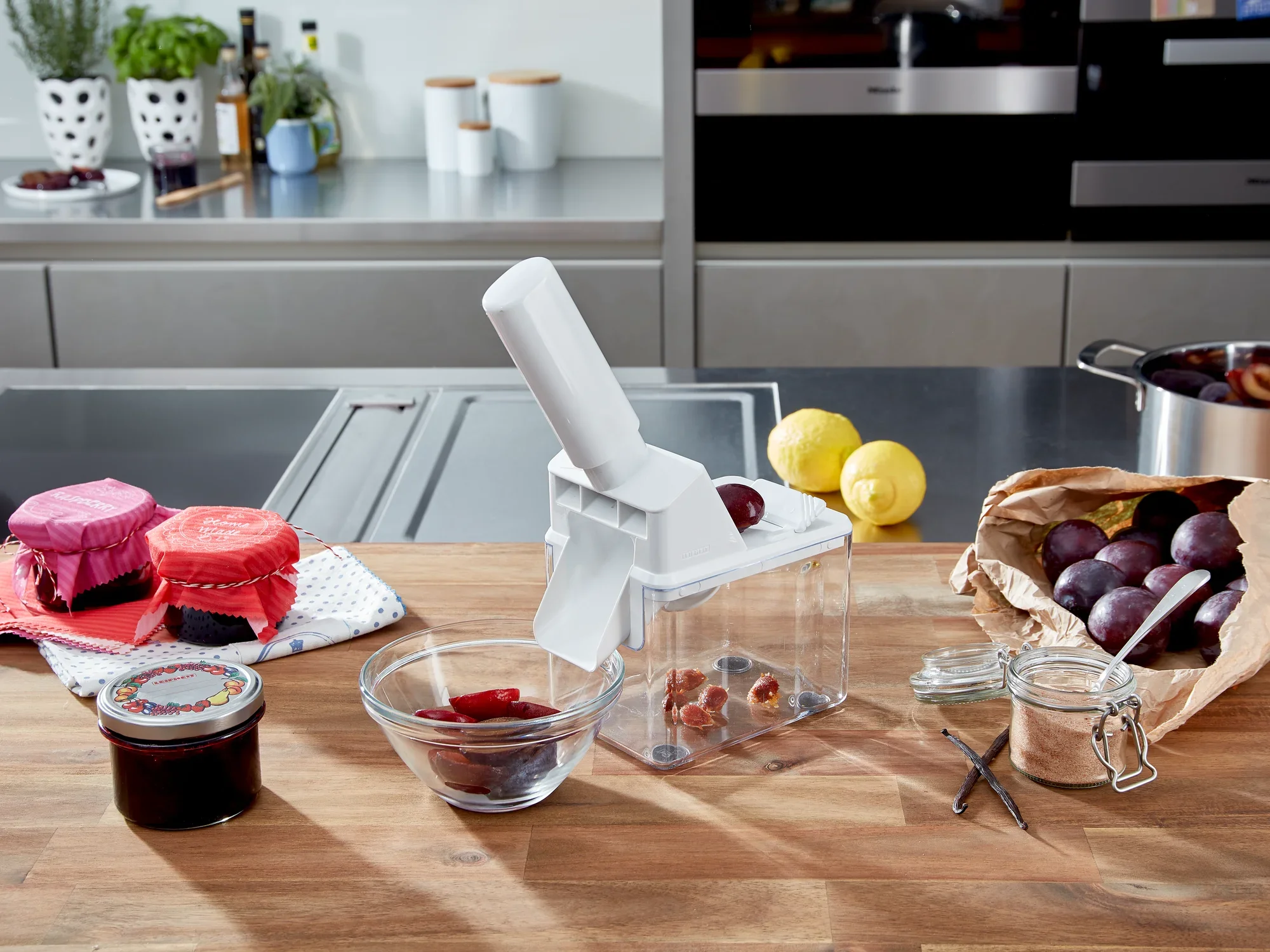 Drylownica do śliwek stoi na nowoczesnym blacie kuchennym. Otoczony świeżymi śliwkami, słoikami z dżemem i przyborami kuchennymi, obraz przedstawia praktyczną scenę kuchenną.