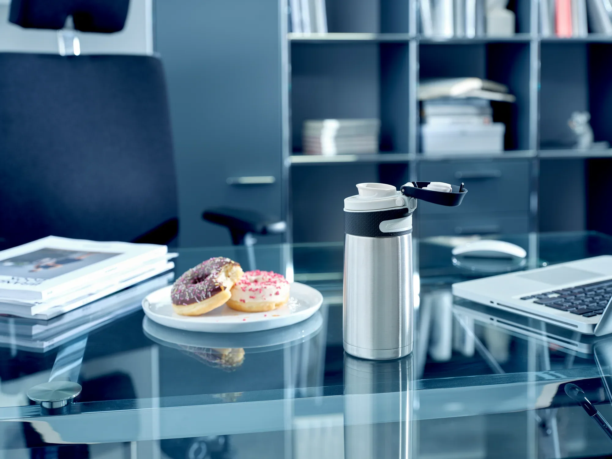 Srebrny kubek próżniowy z pokrywką typu flip o pojemności 350 ml stoi na nowoczesnym szklanym stole biurowym. Obok leżą dwa pączki na talerzu. W tle widoczne są półki z książkami.