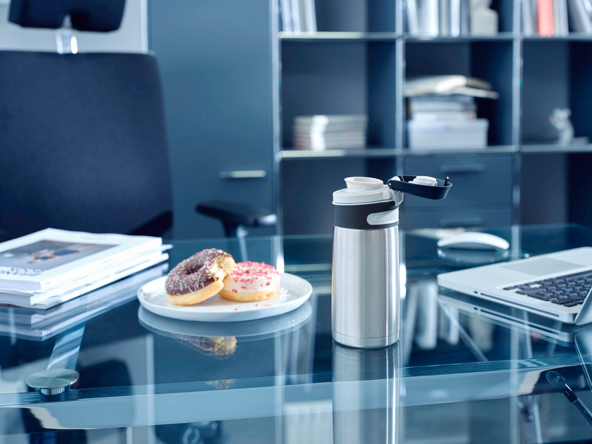 Een zilveren vacuümmok met klapdeksel, 350 ml, staat op een moderne glazen kantoortafel. Ernaast liggen twee donuts op een bord. Op de achtergrond zijn boekenplanken te zien.