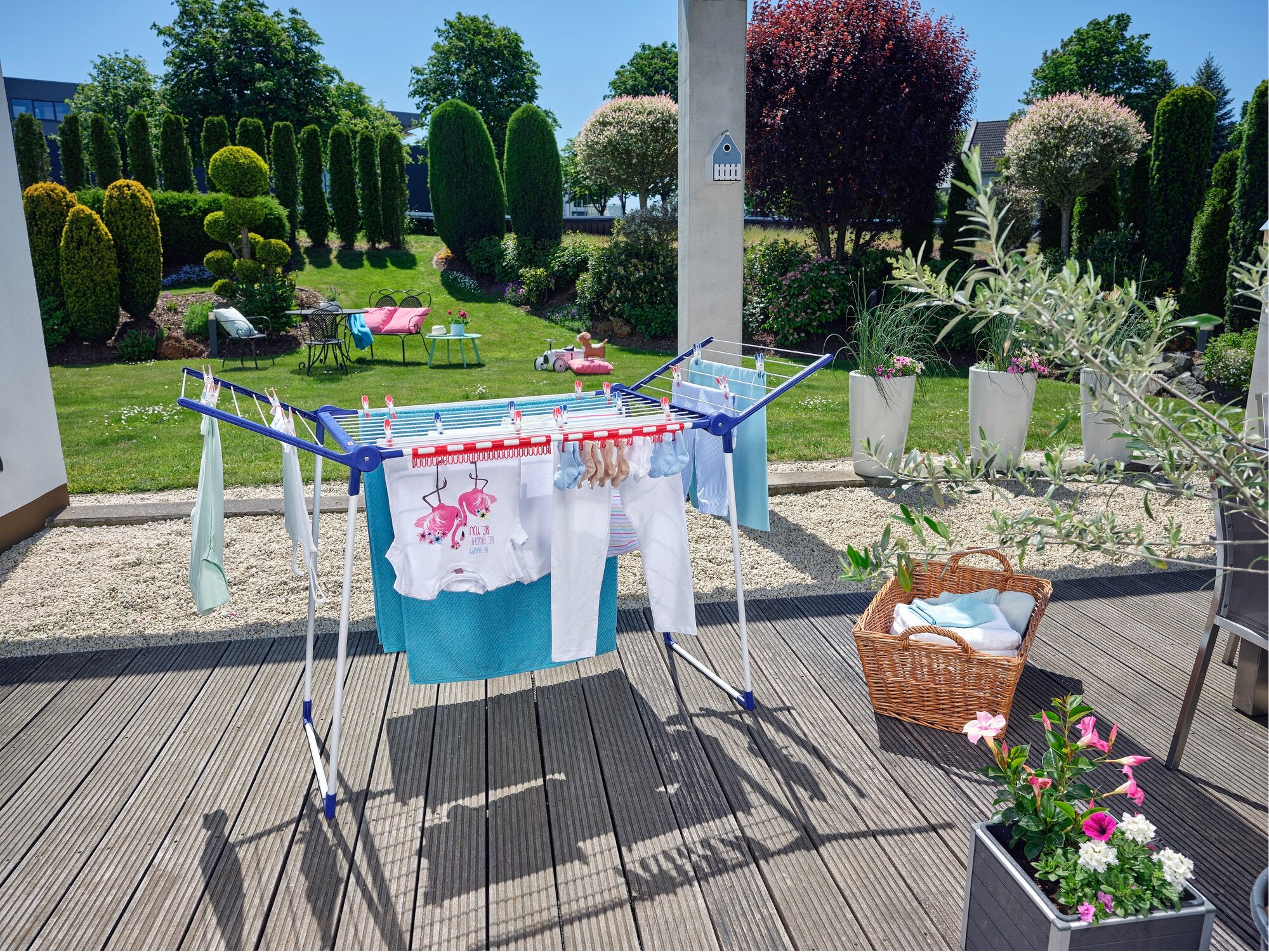 Ein Standtrockner Pegasus 180 Solid Classic steht auf einer Terrasse mit Blick auf einen gepflegten Garten. Auf dem Trockner hängen bunte Kleidungsstücke zum Trocknen.